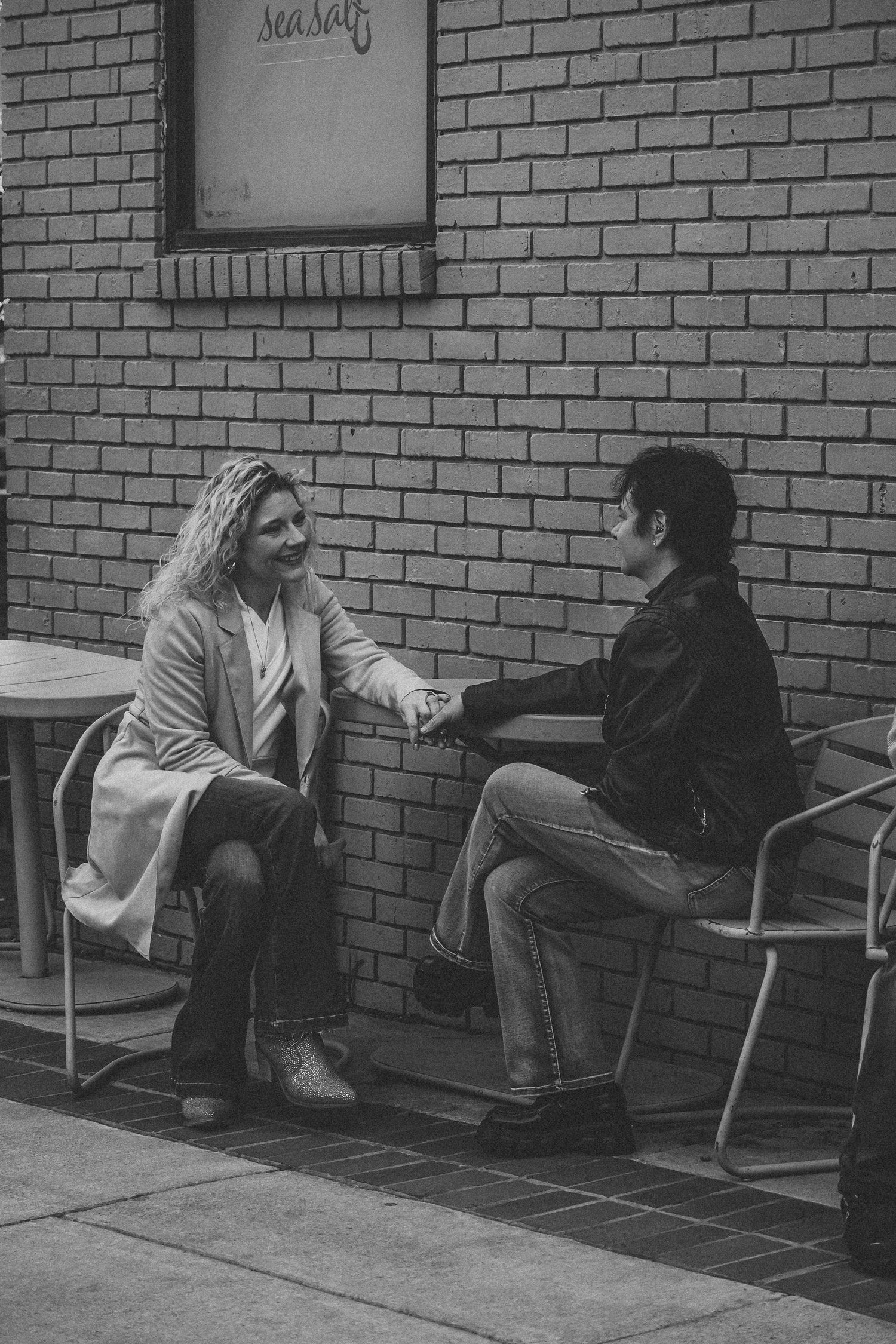 Couple holding hands while seated outside against a brick wall near a Sea Salt sign in black and white.