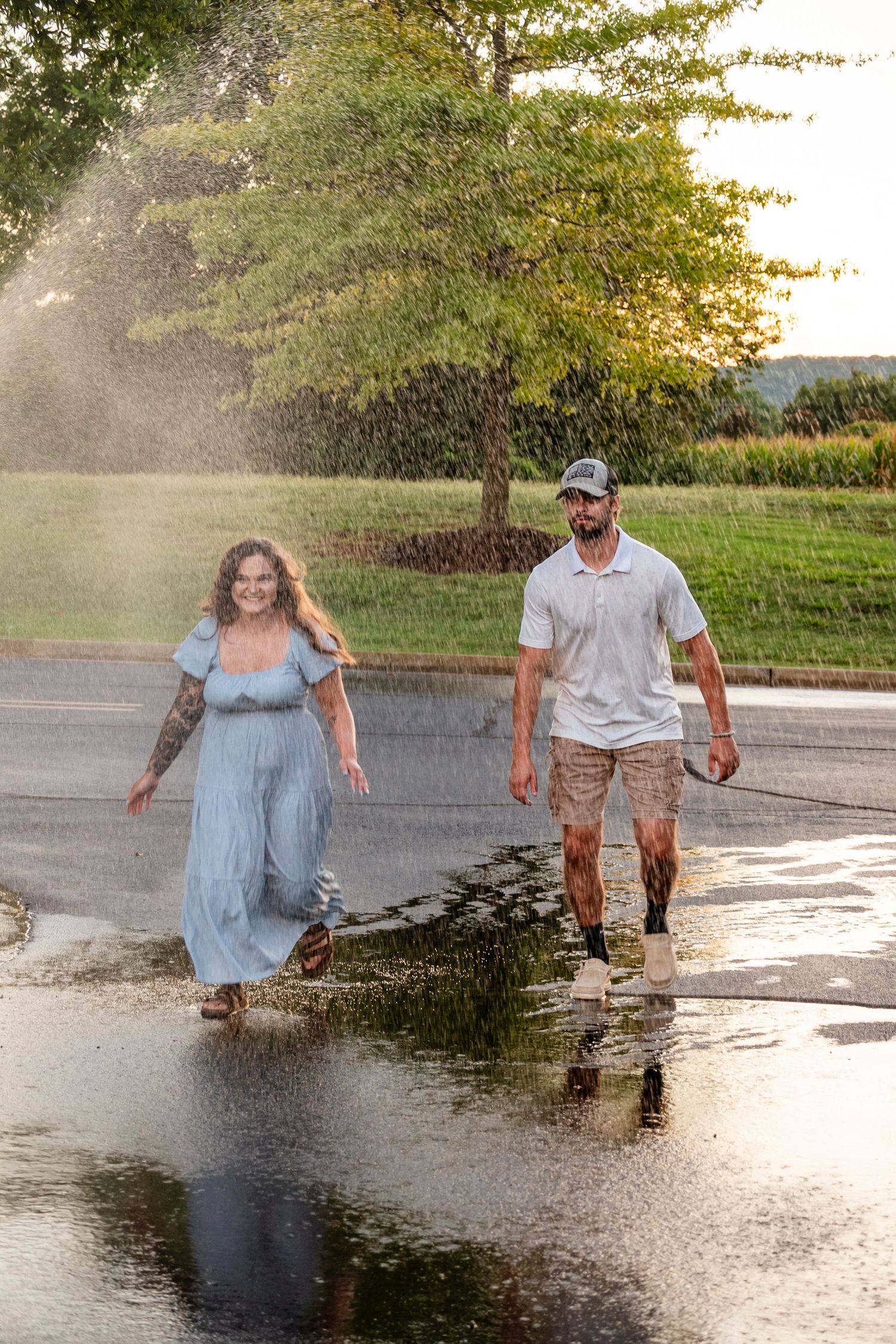 A smiling couple walks through a sprinkler on a wet road surrounded by lush green trees.