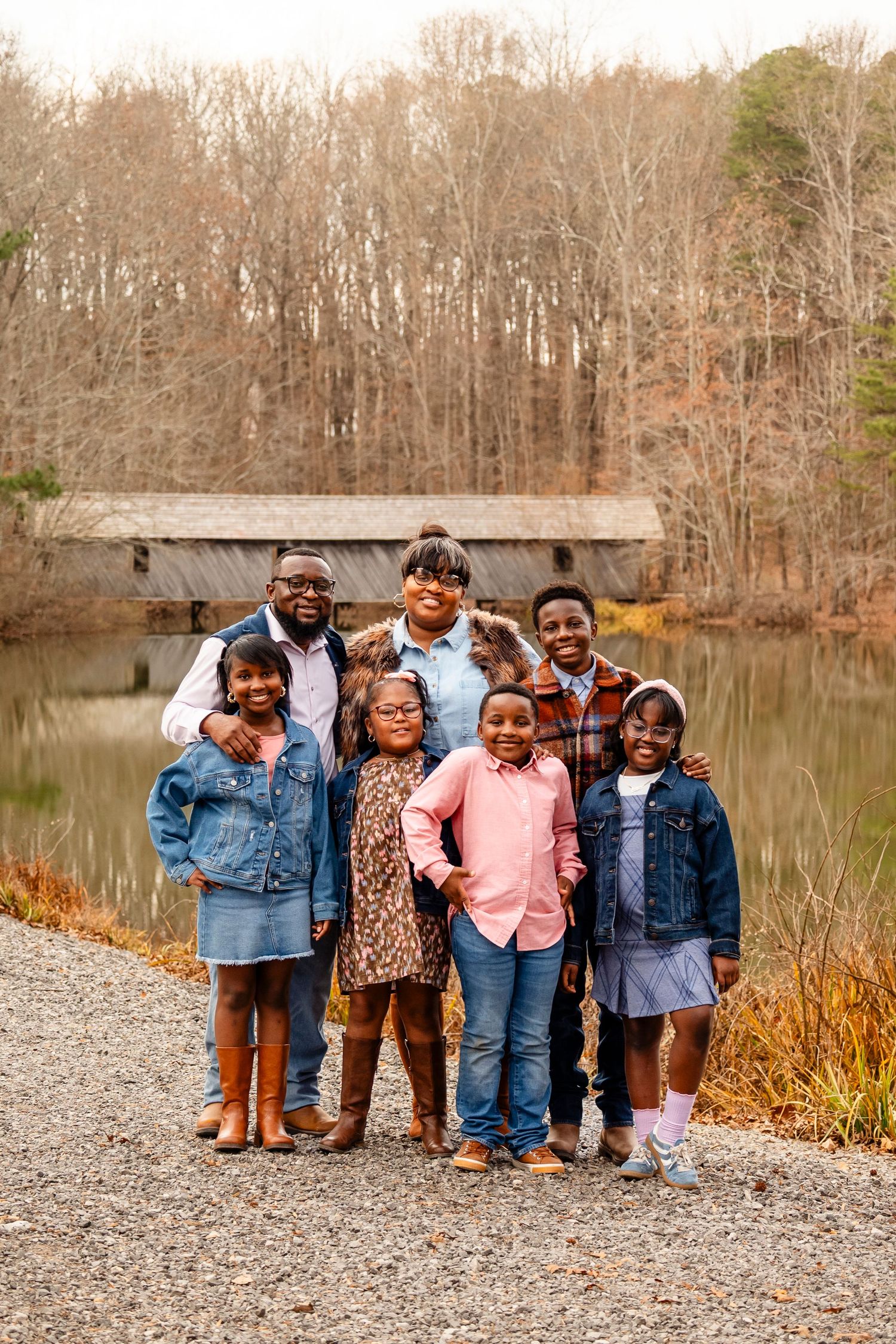 Family of seven posing together near a pond with a covered bridge in autumn woods.