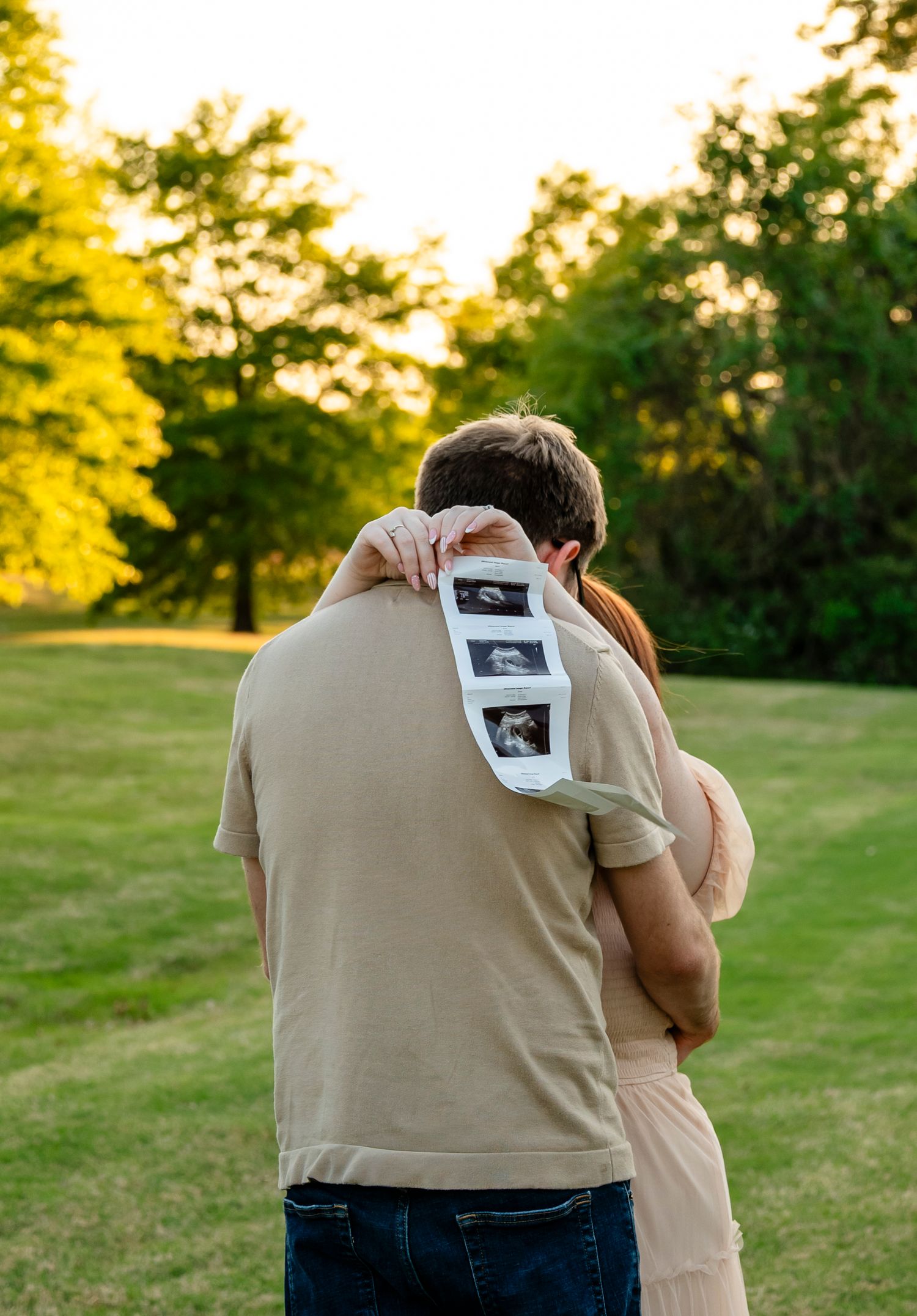 Couple embracing outdoors holding ultrasound photos, pregnancy announcement in a lush green park at golden hour.