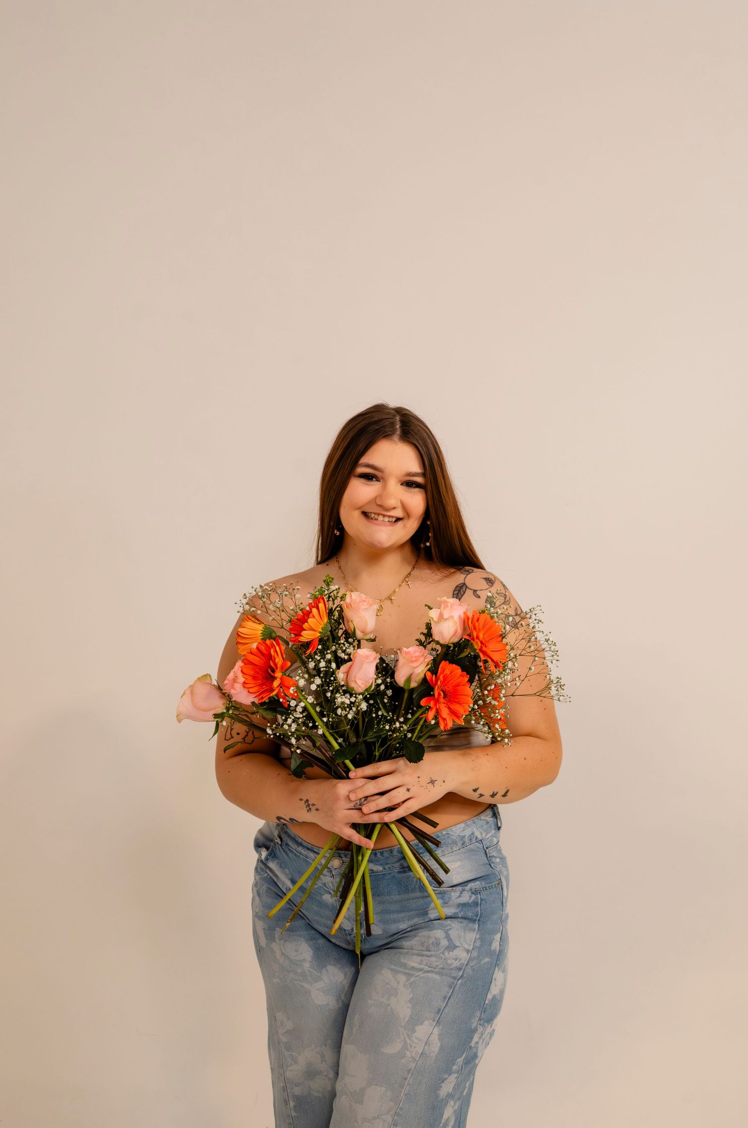 Smiling brunette woman in floral top and jeans holds a bright bouquet of orange and pink flowers.