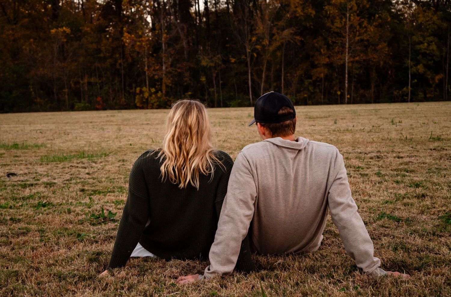 Couple sitting together on dry grass field, viewed from behind with autumn trees in background.