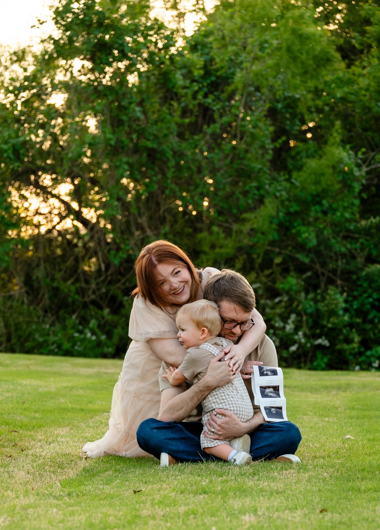 Happy family maternity announcement on grass with parents hugging toddler holding ultrasound photos at golden hour.