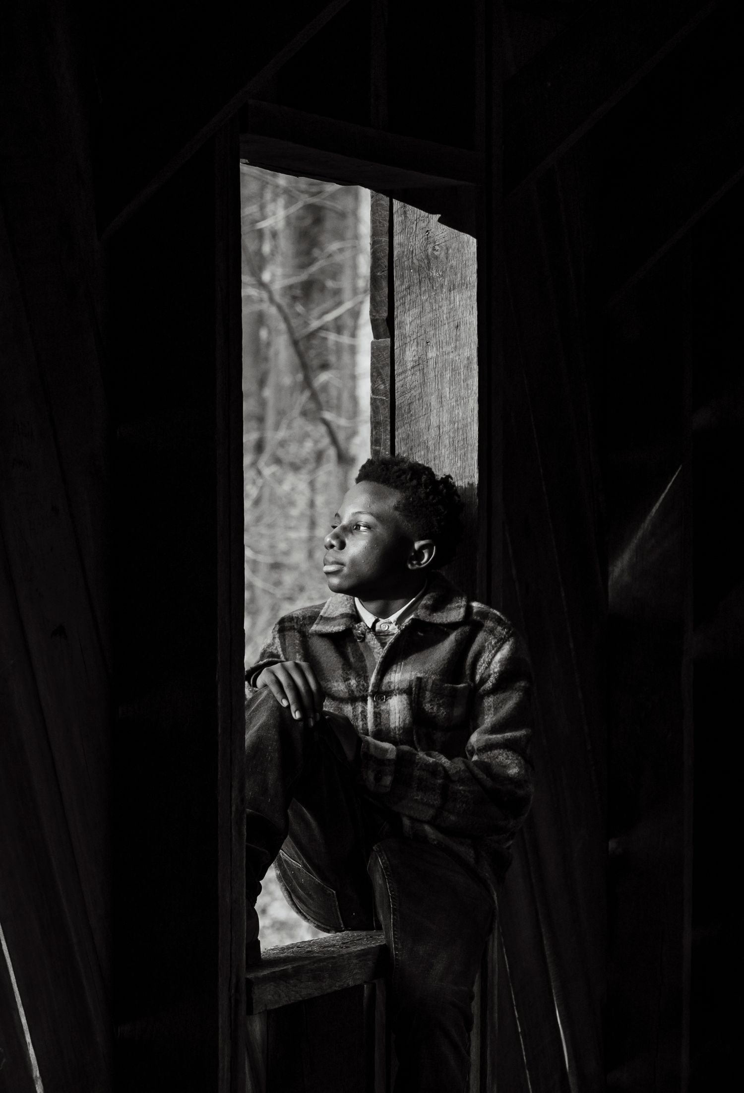 A black and white portrait of a young Black boy gazing out a dark wooden cabin doorway.