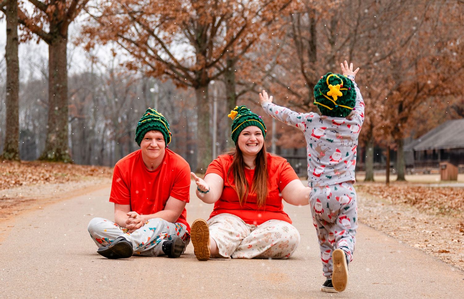 Family in matching red outfits and green beanies sitting on path as toddler runs toward them in autumn park.
