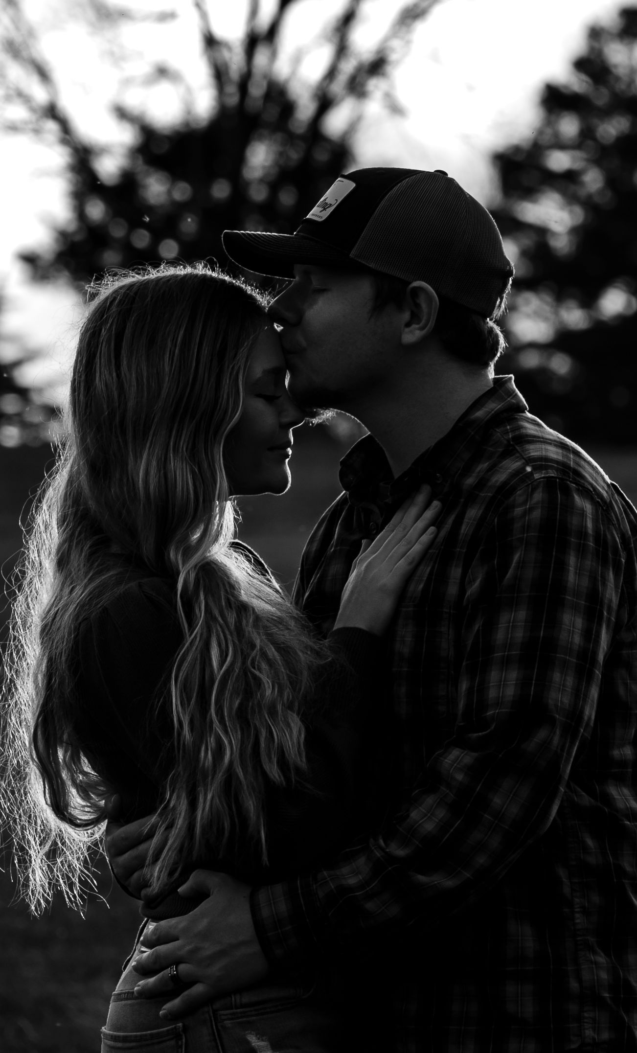 Black and white silhouette of a couple about to kiss at dusk beneath dark trees.