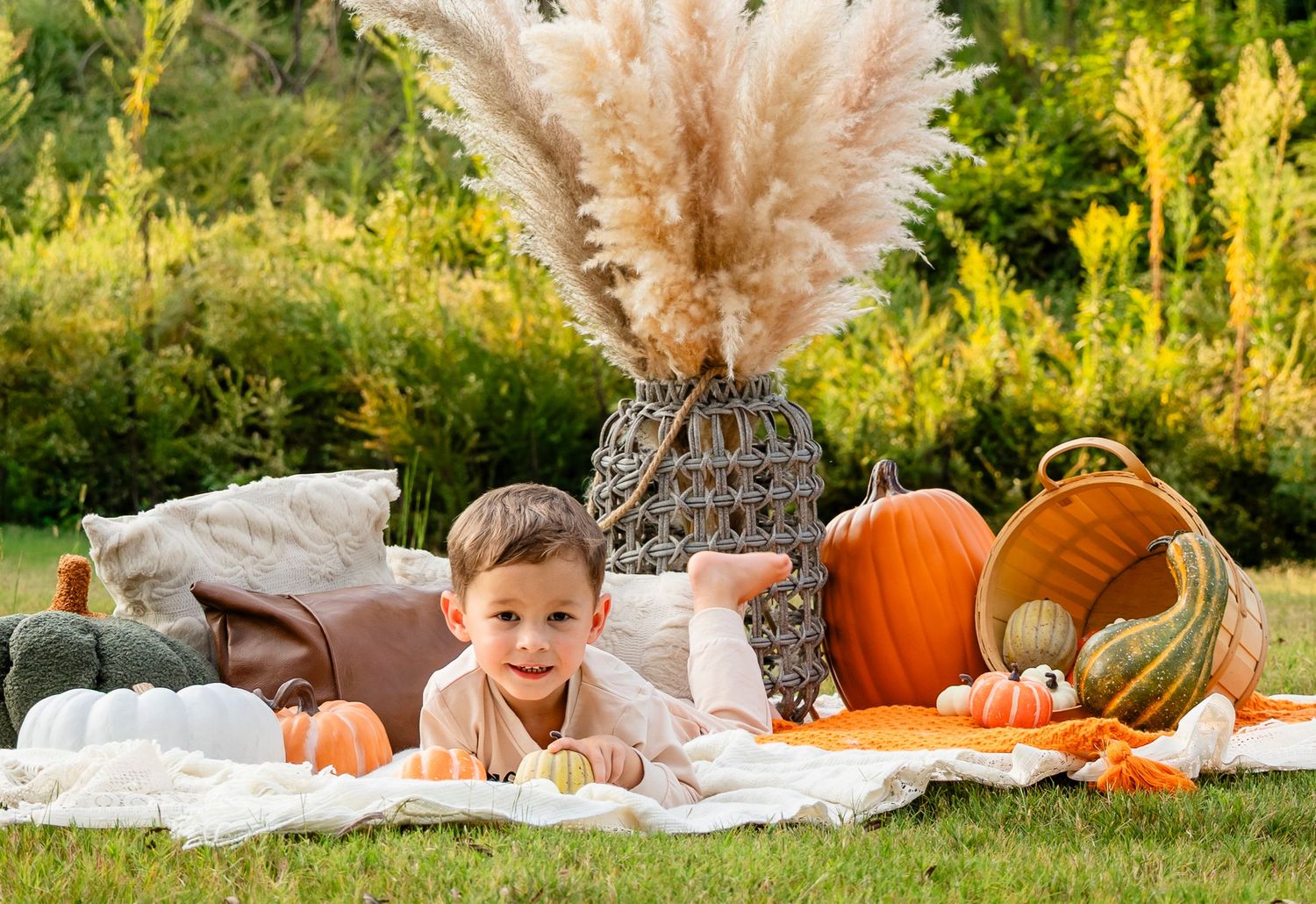 Young boy lying on white blanket surrounded by pumpkins and fall decor in sunny garden.