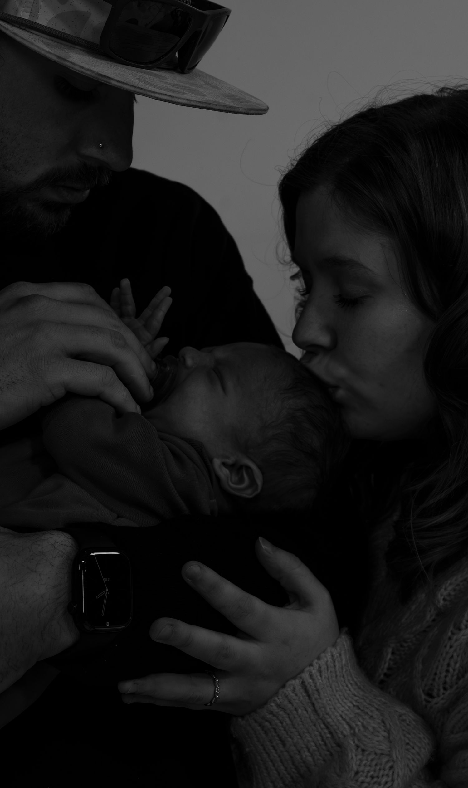 Dark intimate photo of parents sharing a tender moment kissing their newborn baby.