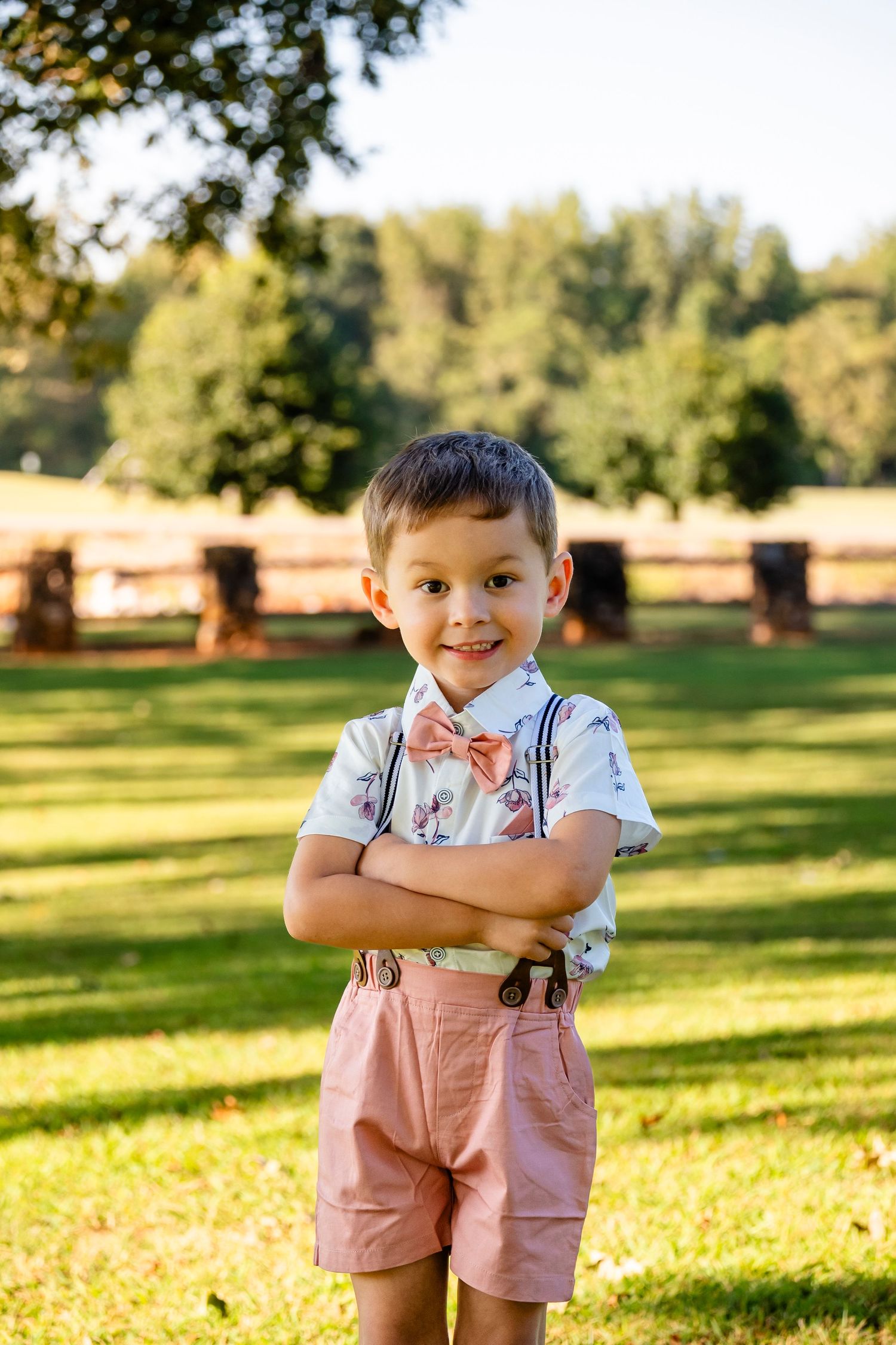 Adorable young boy in pink bow tie, suspenders, and shorts stands with arms crossed smiling in sunny park.