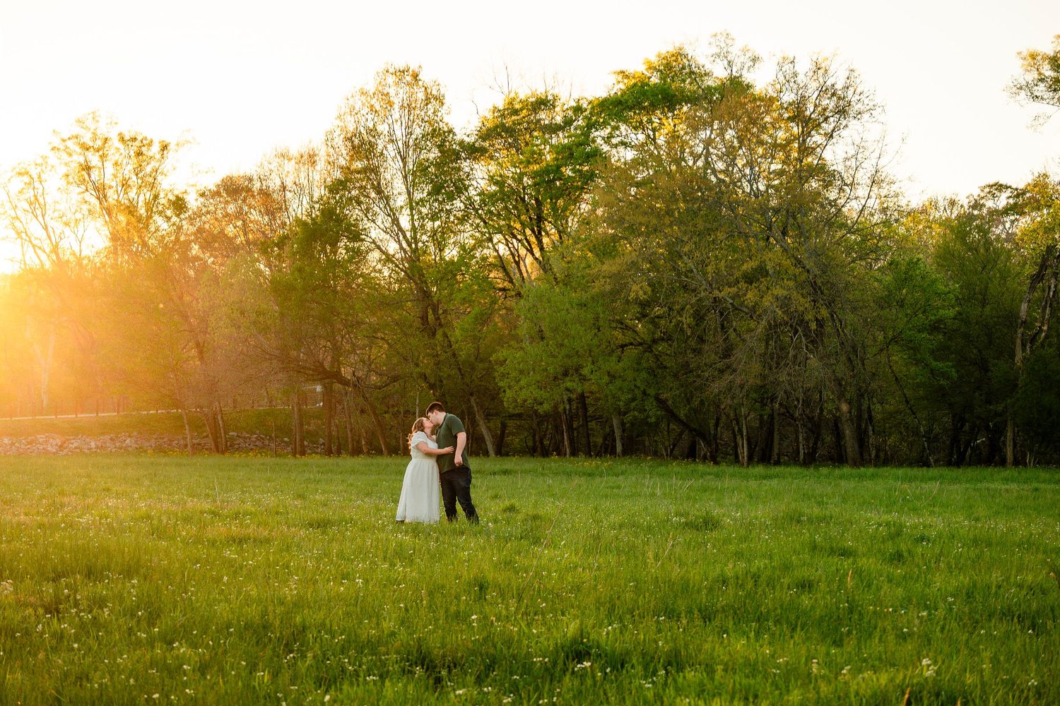 Couple embracing in a sunlit green meadow at golden hour with lush trees in the background at a wedding.