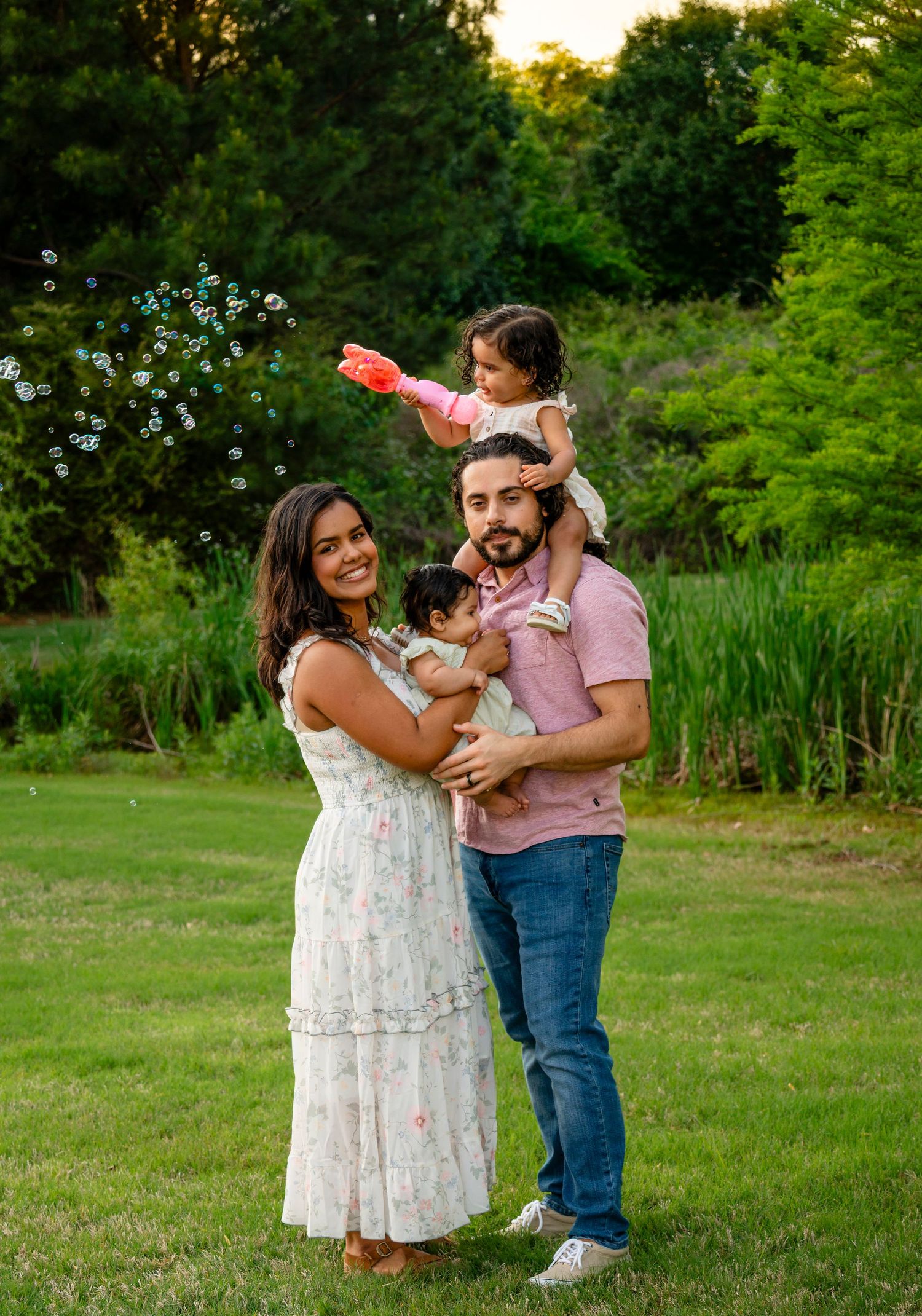 Family of four posing outdoors on green lawn, mother in floral dress, father holding toddler blowing bubbles at golden hour.