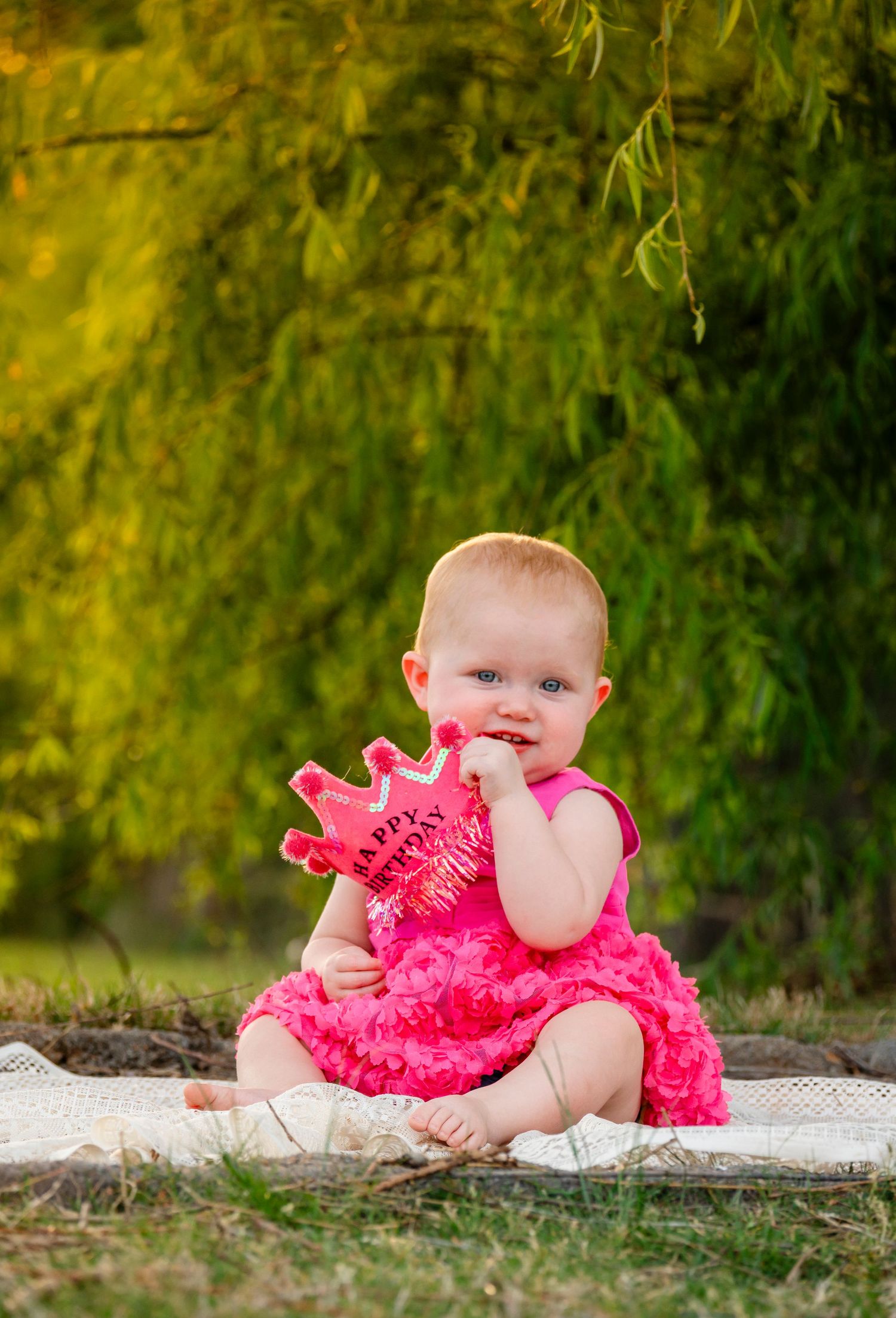 Cute baby girl in pink ruffled dress sits on blanket holding a Happy Birthday crown prop.