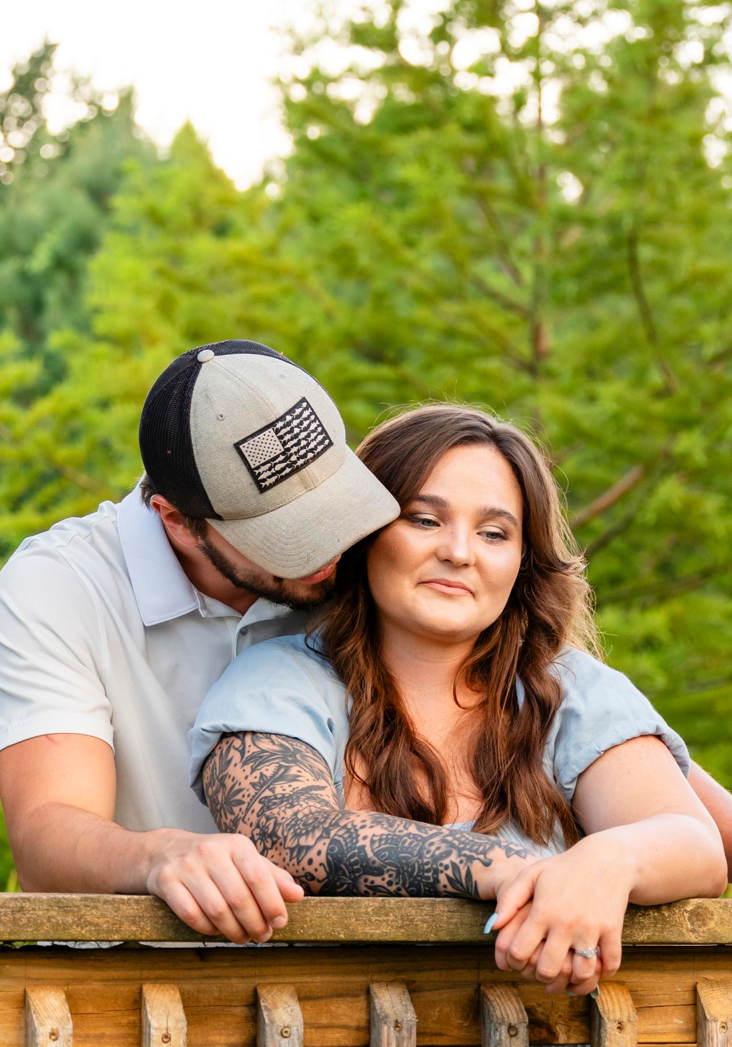 A tattooed man in a cap kisses a smiling woman's cheek while leaning on a rustic wooden railing.