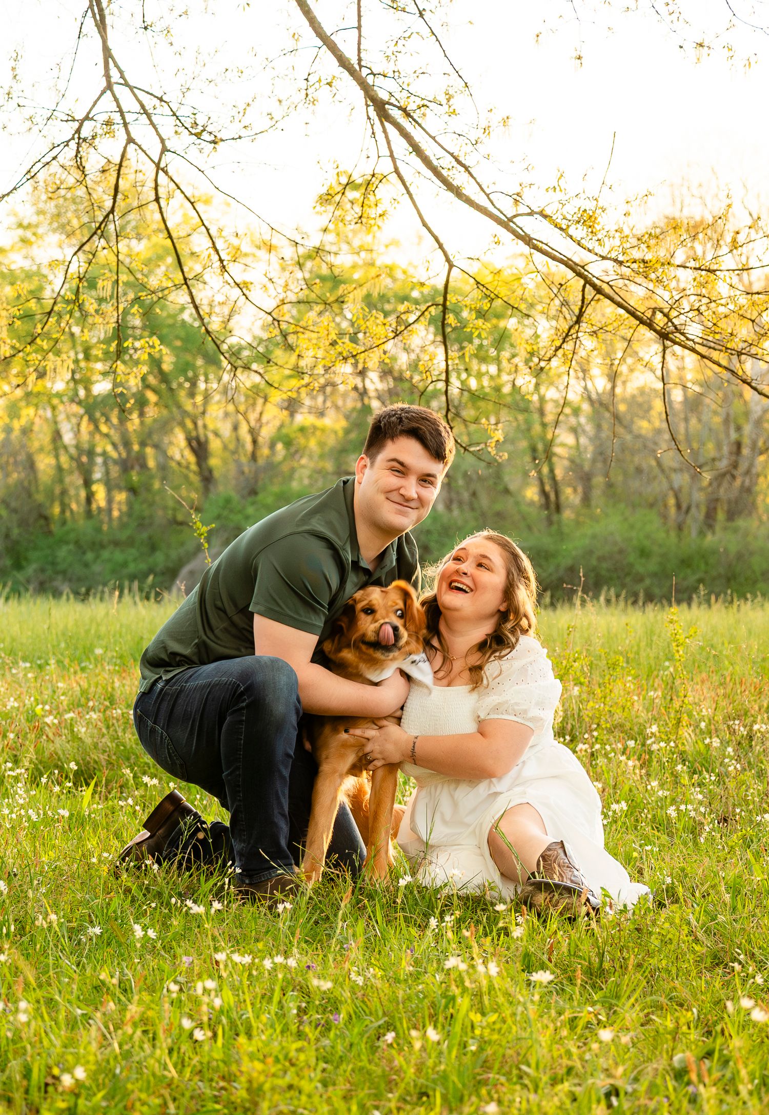 Happy couple laughing and cuddling with their golden dog in a sunny green meadow at sunset.