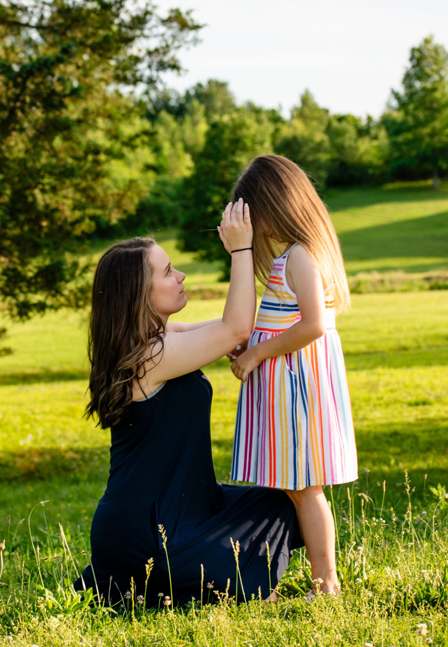 A mother kneels in a sunny green field, gently brushing hair from her young daughters face outdoors.