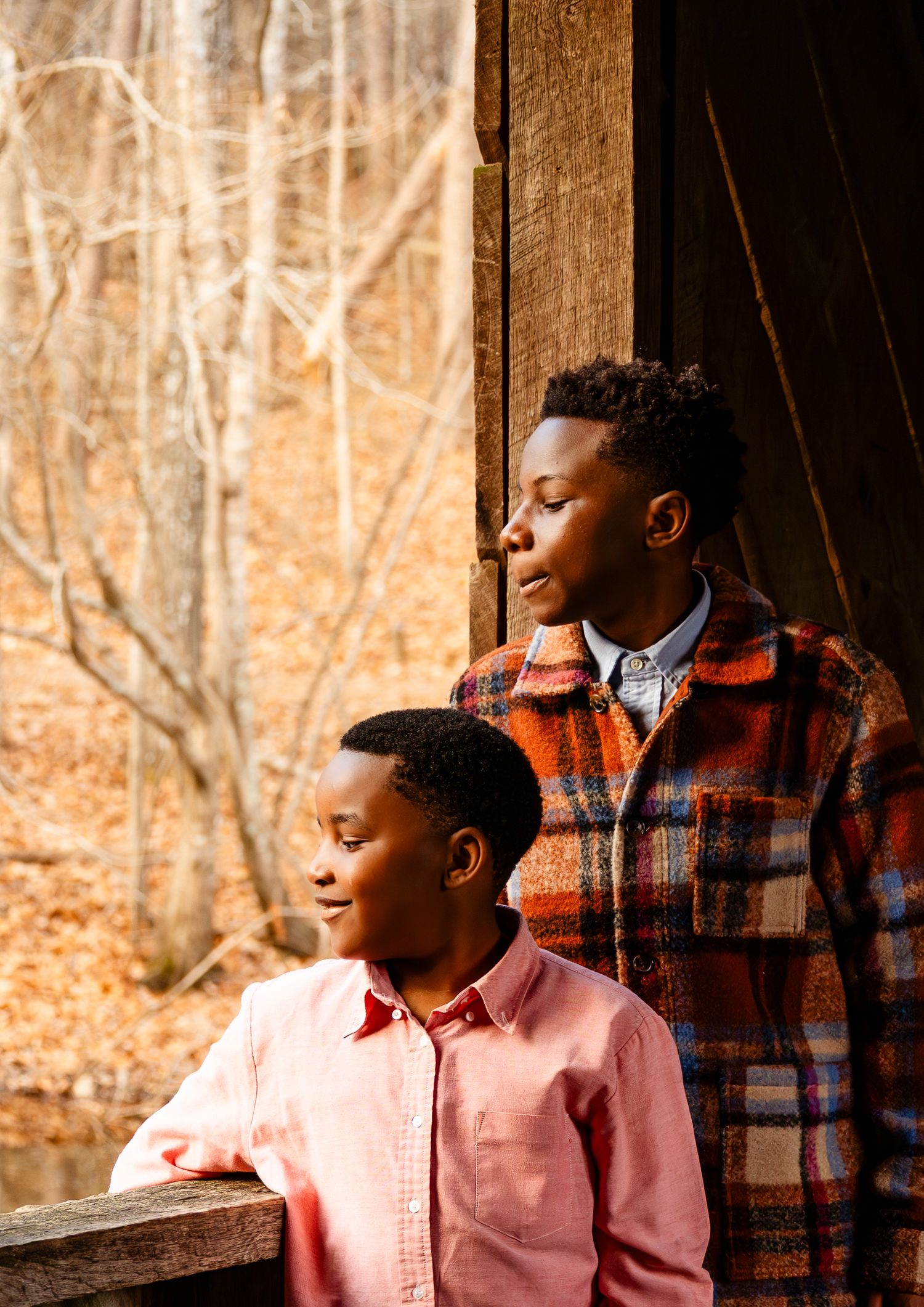 Two Black boys gaze thoughtfully out a rustic wooden cabin window surrounded by fall foliage.