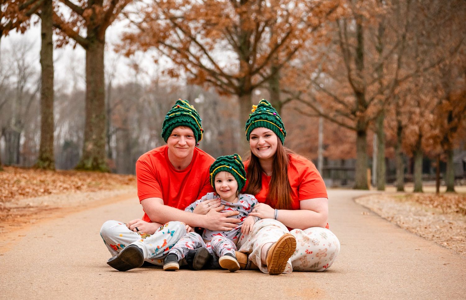 Family of three wearing green Christmas tree hats sits together on a leaf-covered outdoor path.