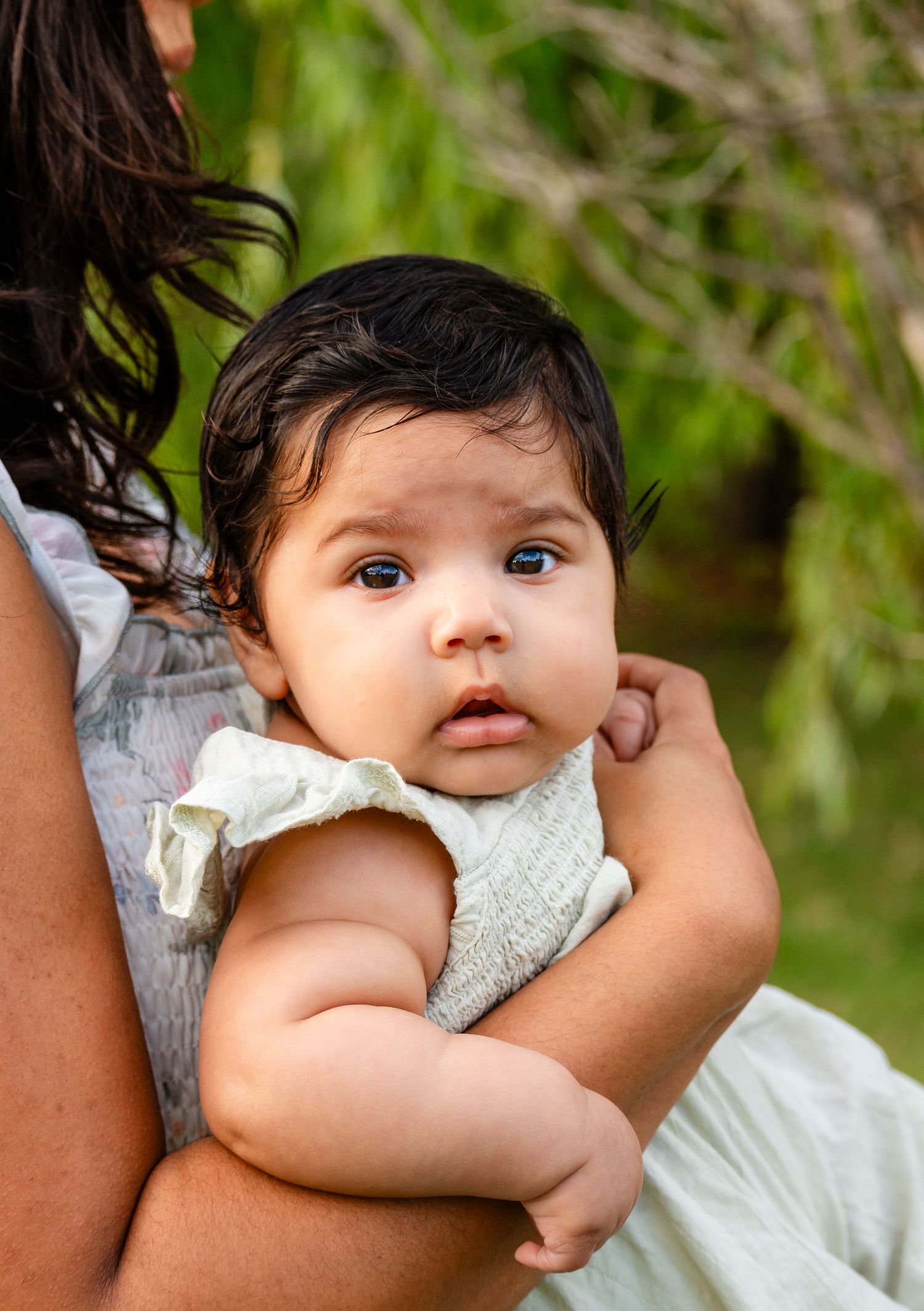 A dark-haired baby in a white romper gazes curiously while being held outdoors near green trees.