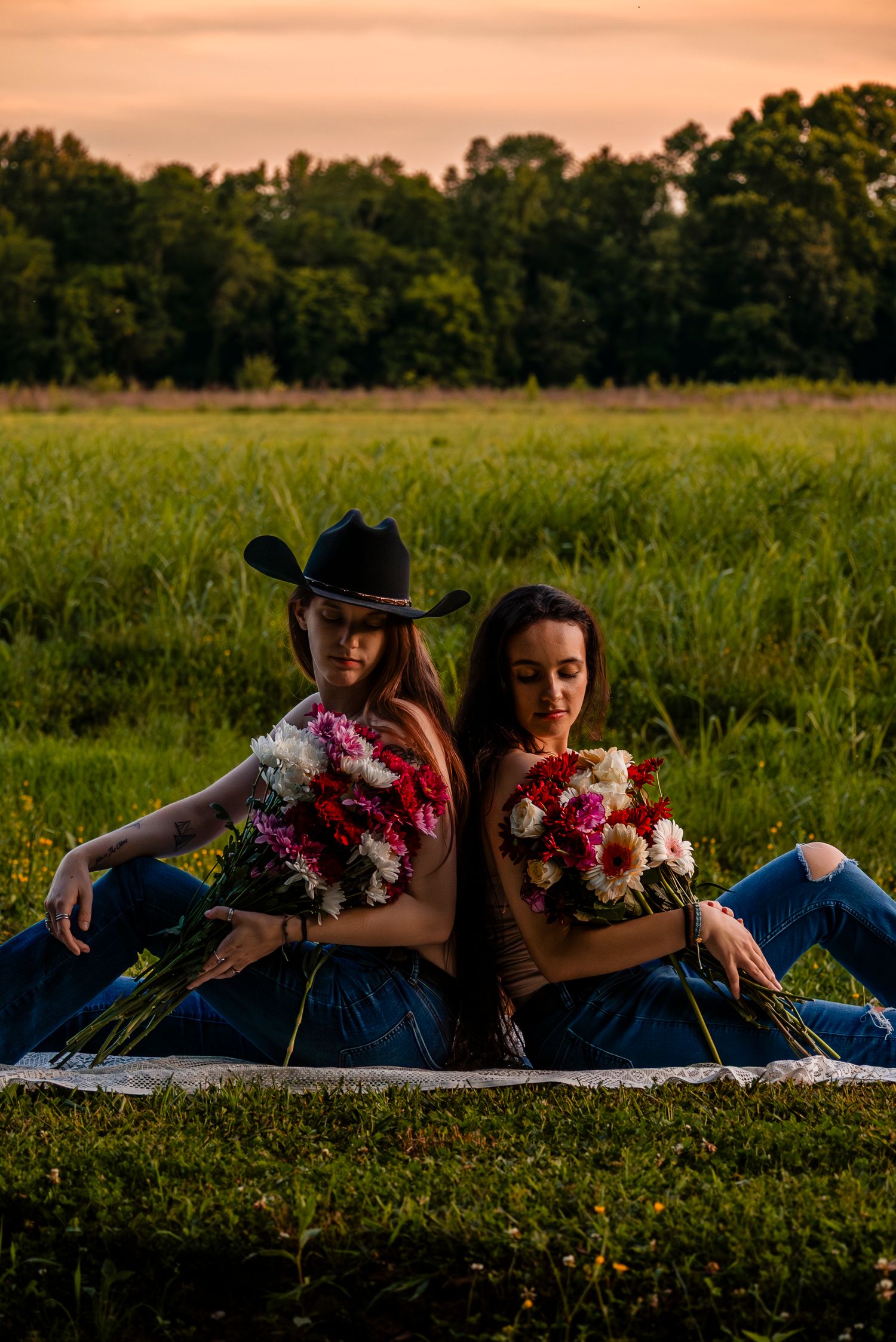 Two young women sit back-to-back in a field at sunset, holding colorful flower bouquets, one wearing a black cowboy hat.