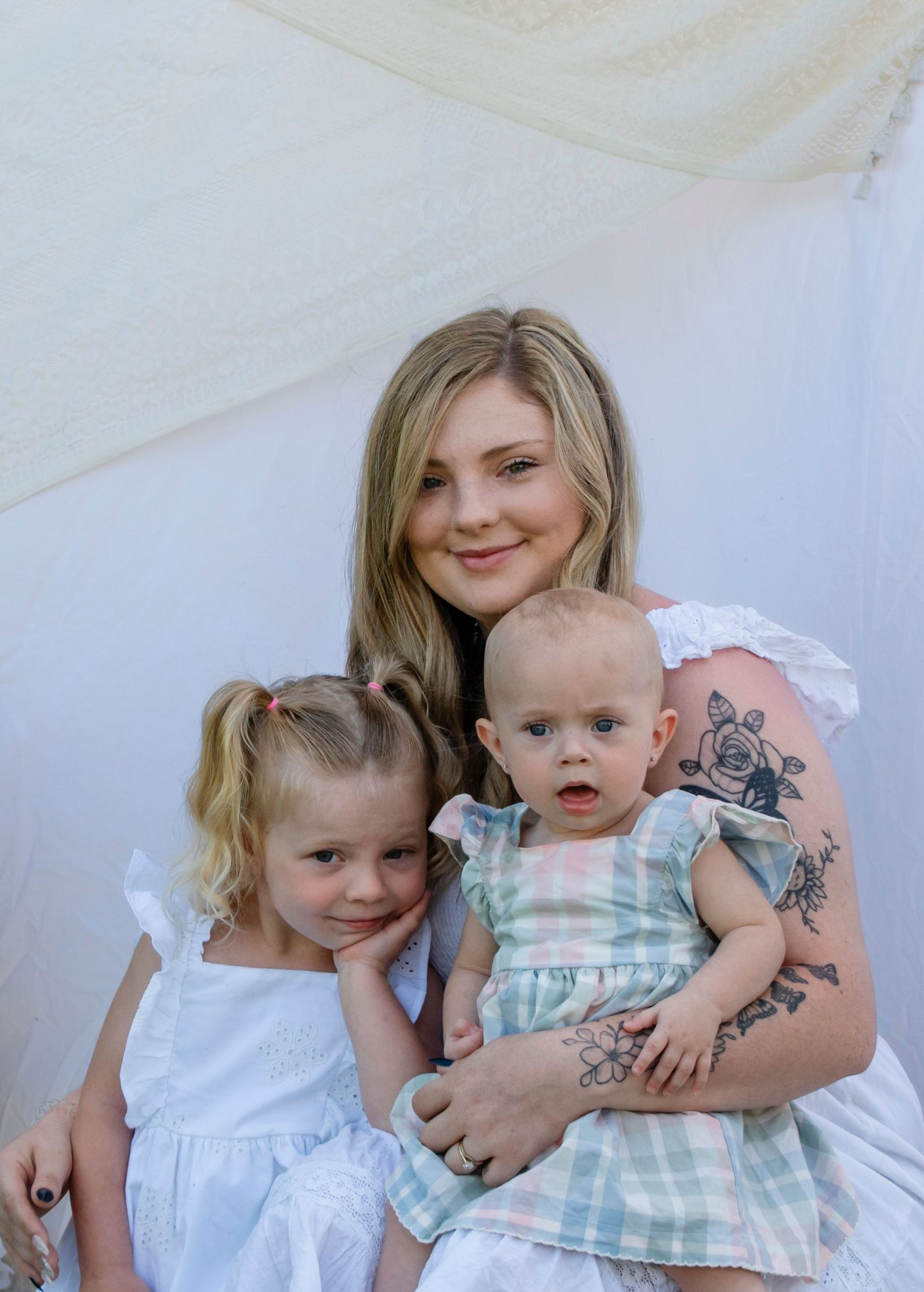 A young blonde mother with a floral tattoo smiles while holding her toddler and infant daughters in white outfits.