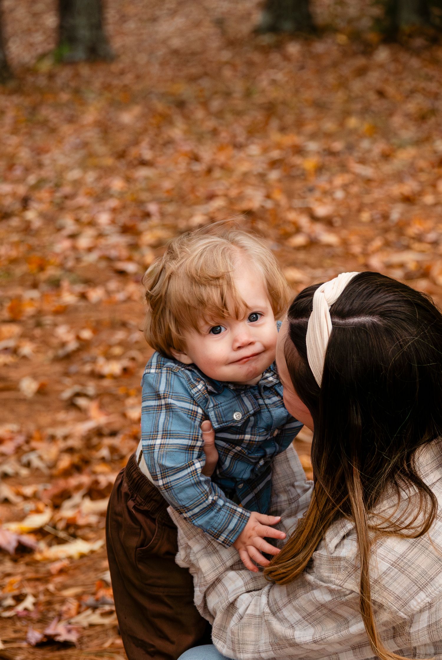 Mother kissing toddler's cheek surrounded by autumn leaves in wooded park.