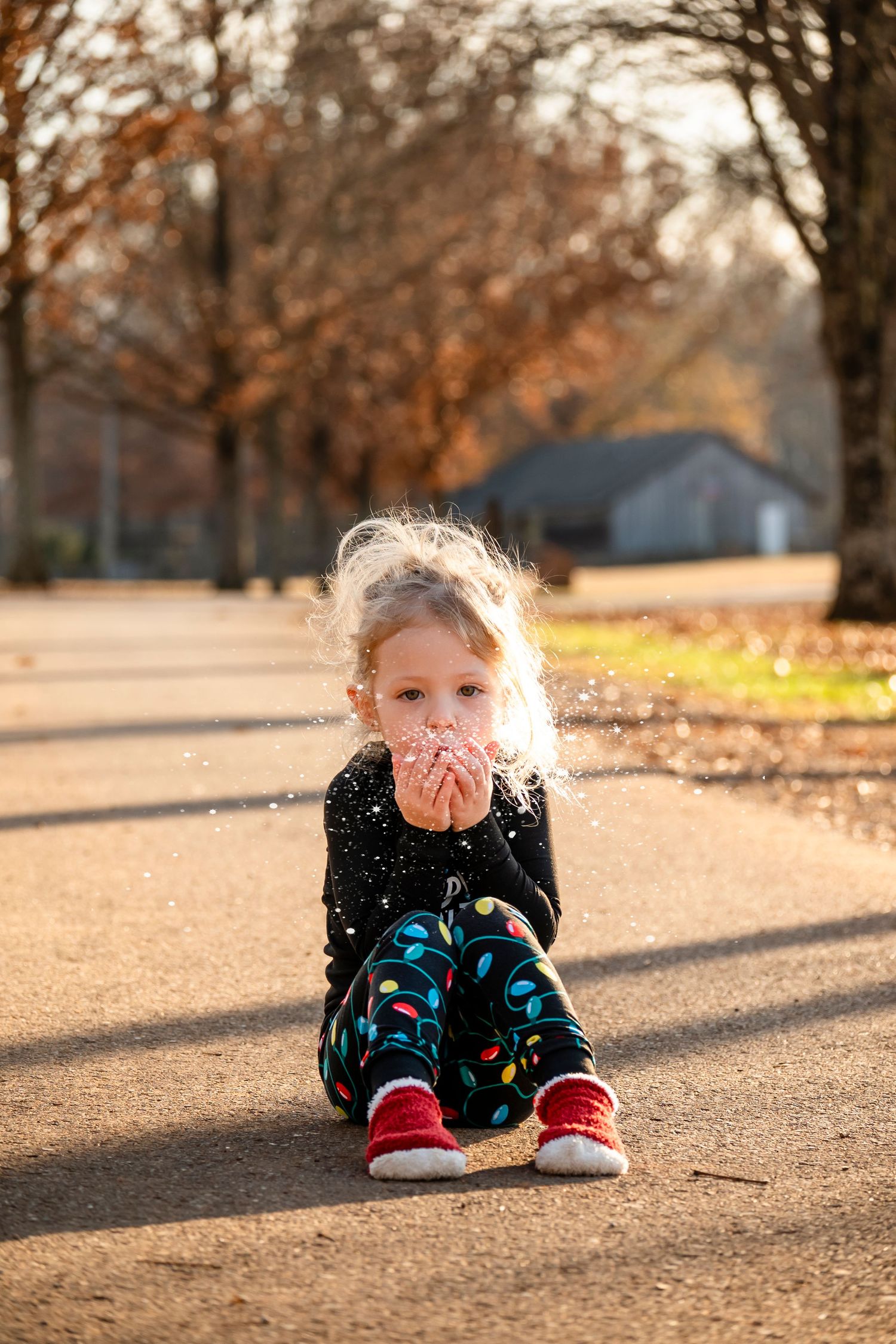 Cute blonde toddler in Christmas light pajamas blowing fake snow while sitting on outdoor path.