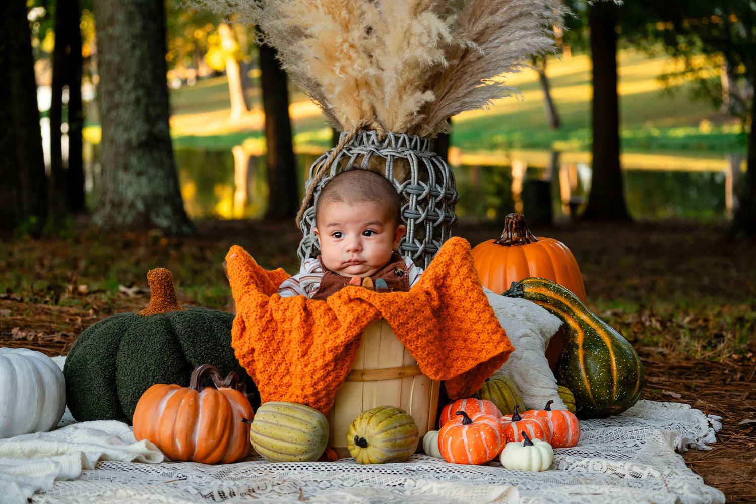 Baby in orange knit wrap sitting in wooden barrel among colorful pumpkins in autumn garden.
