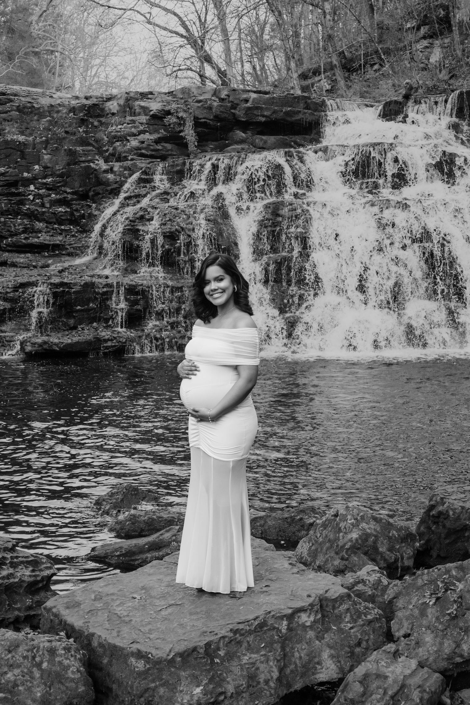 Pregnant woman in white off-shoulder gown standing on rocks before a cascading waterfall, cradling her baby bump.