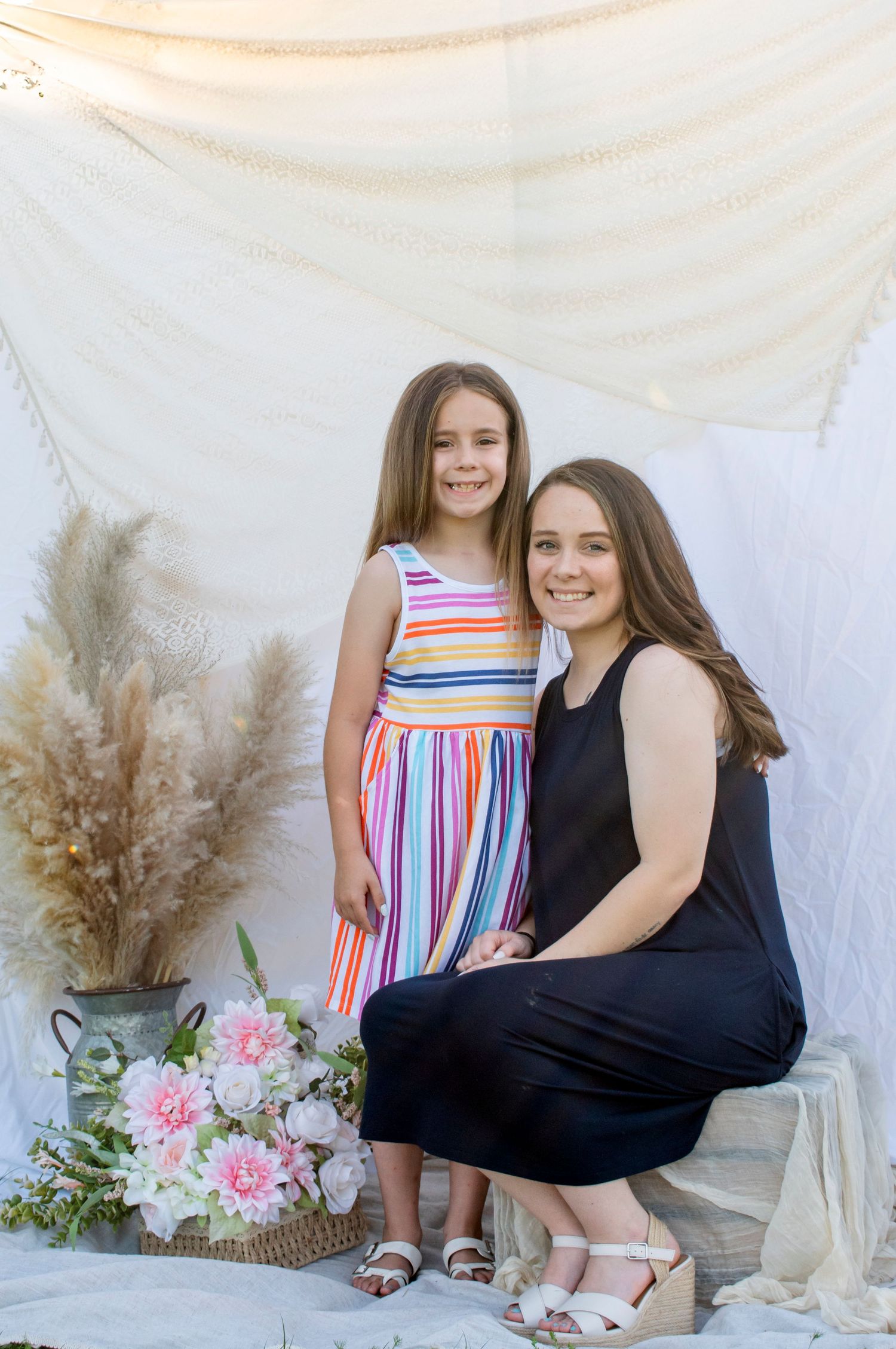 A woman in a black dress crouches beside a young girl in a rainbow striped dress during an outdoor photo session.