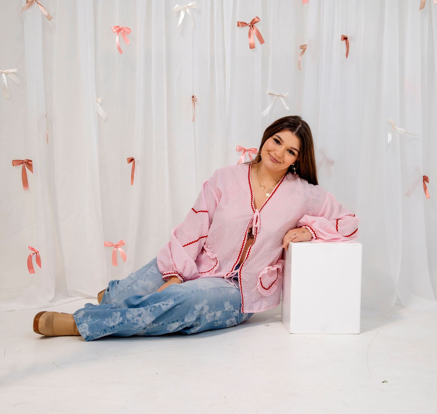 Young woman in pink cardigan and jeans sits on white cube against a white backdrop with pink bows.