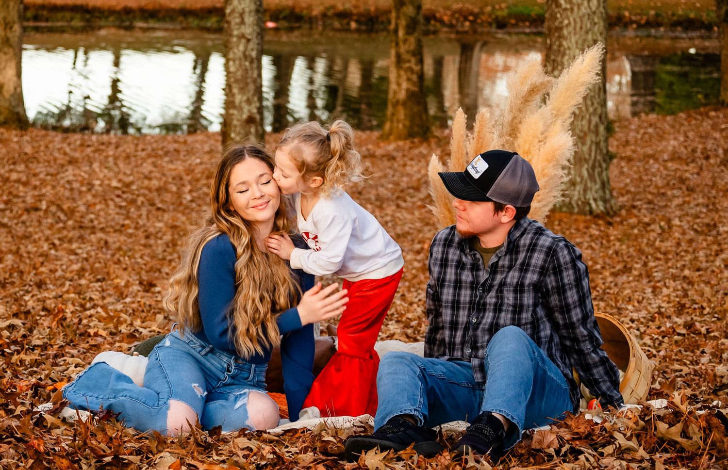 Toddler girl kisses mom while dad smiles during fall family portrait session in autumn leaves.