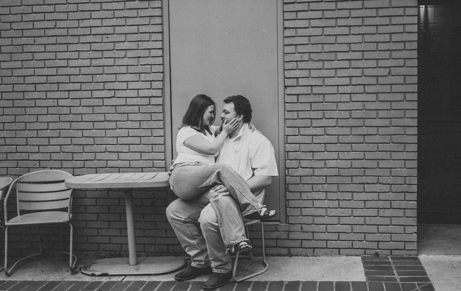 Pregnant woman and partner sharing tender moment seated against brick wall in black and white maternity photo.