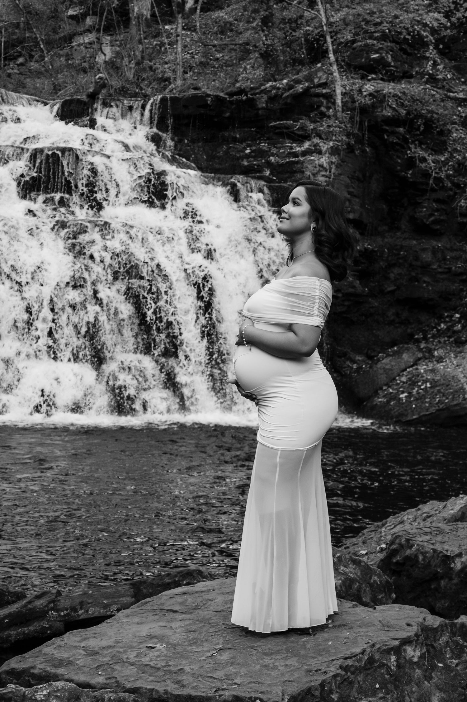 A pregnant woman in a white gown stands gracefully before a waterfall in a black and white photo.