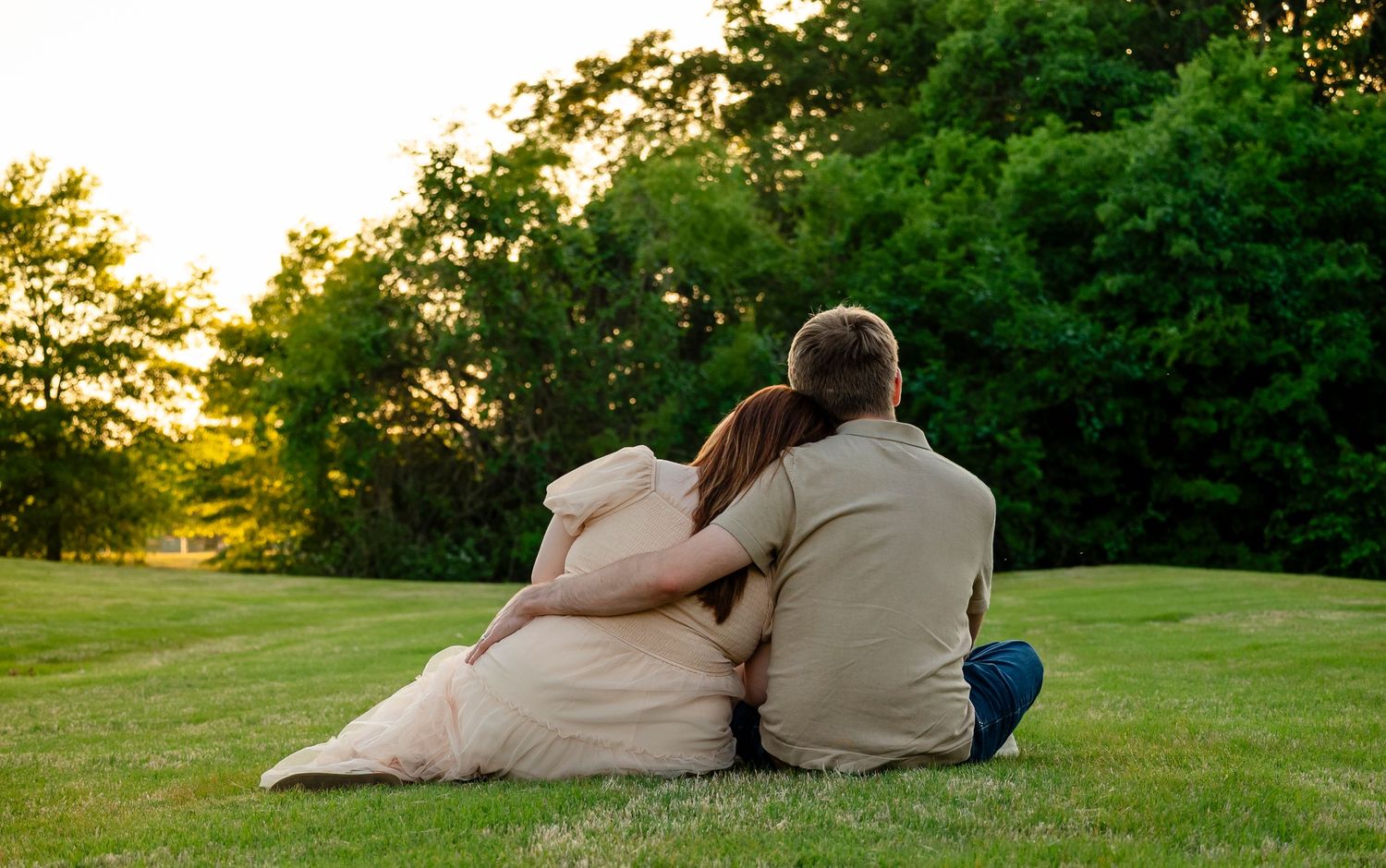 Couple sitting together on green grass at sunset, embracing with lush trees in the background.