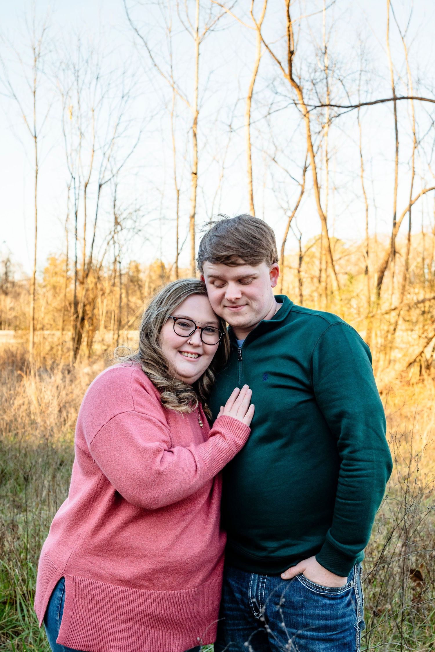 Couple embracing in a golden field at sunset, woman in pink sweater smiling, man in green sweater, surrounded by bare trees.