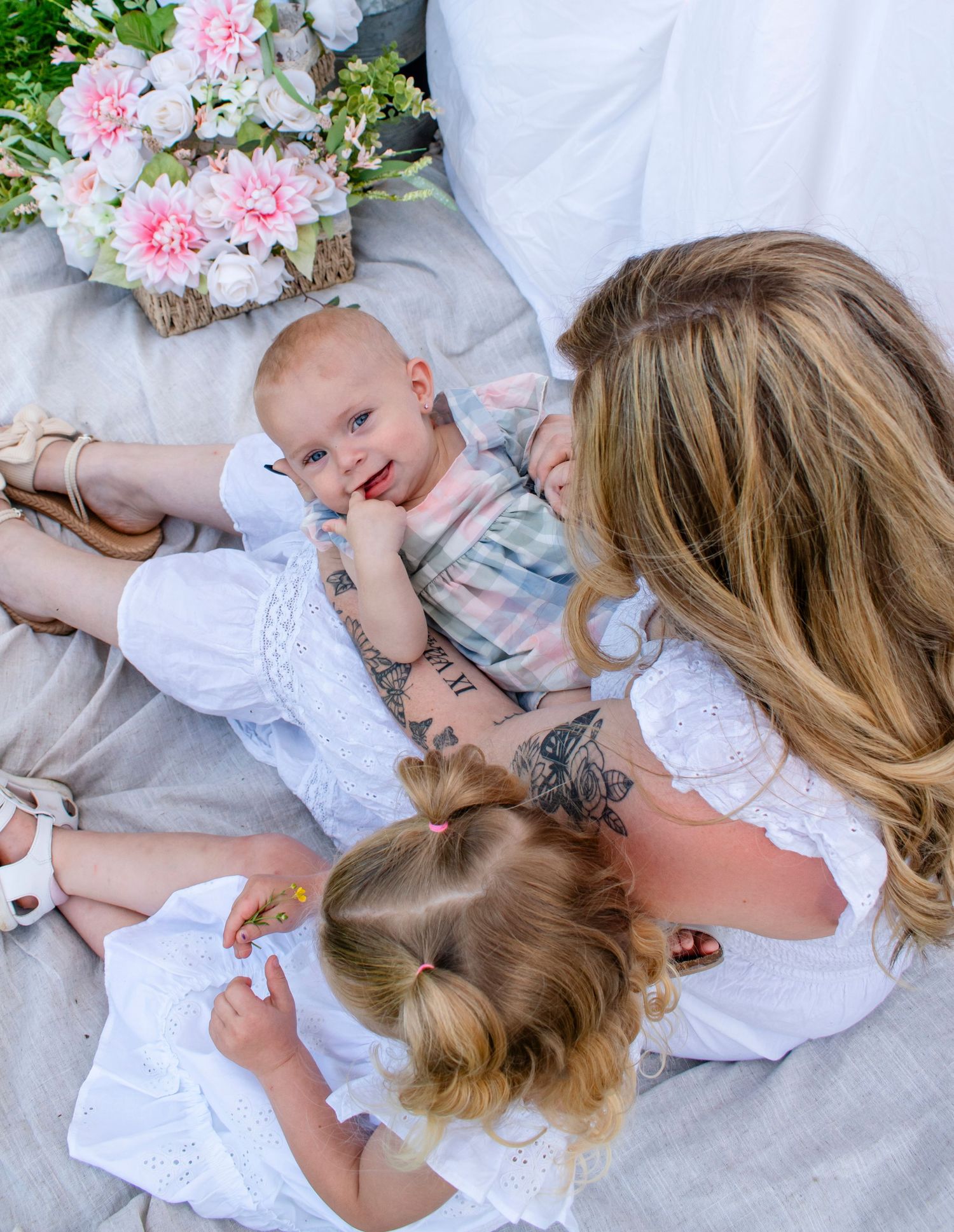 Aerial view of mother with tattoos cuddling two young children on a light blanket with pink flowers nearby.