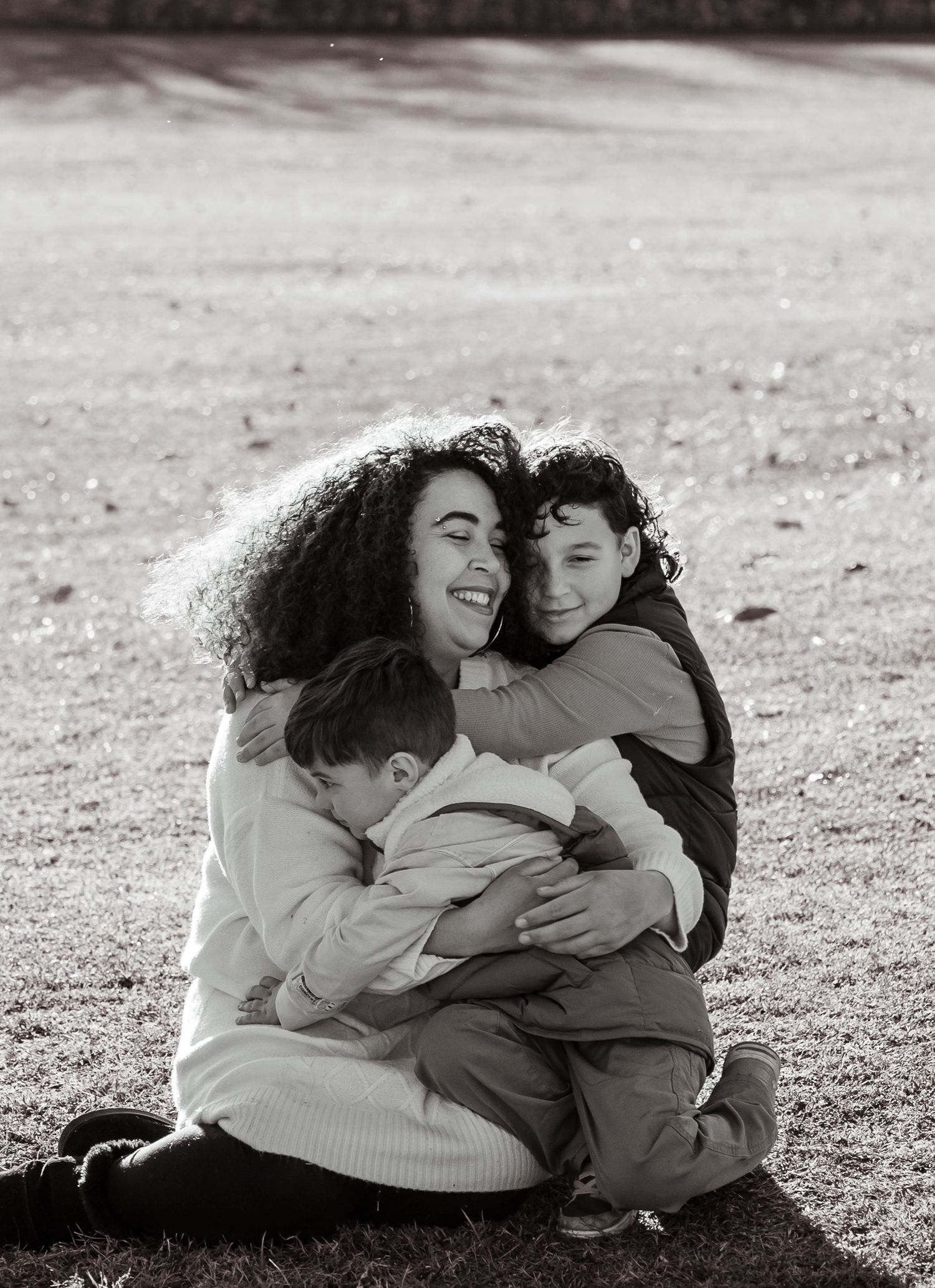 Black and white photo of a laughing mother embracing her two children while sitting on a grassy field.