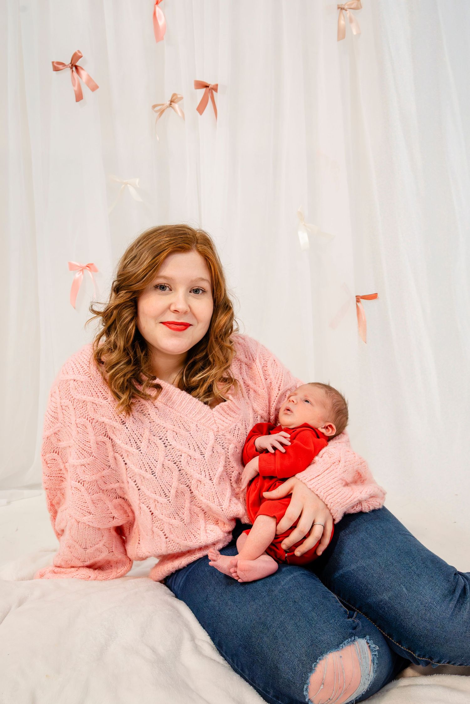 Smiling young mother in pink knit sweater holds her newborn baby in red outfit against bow backdrop.