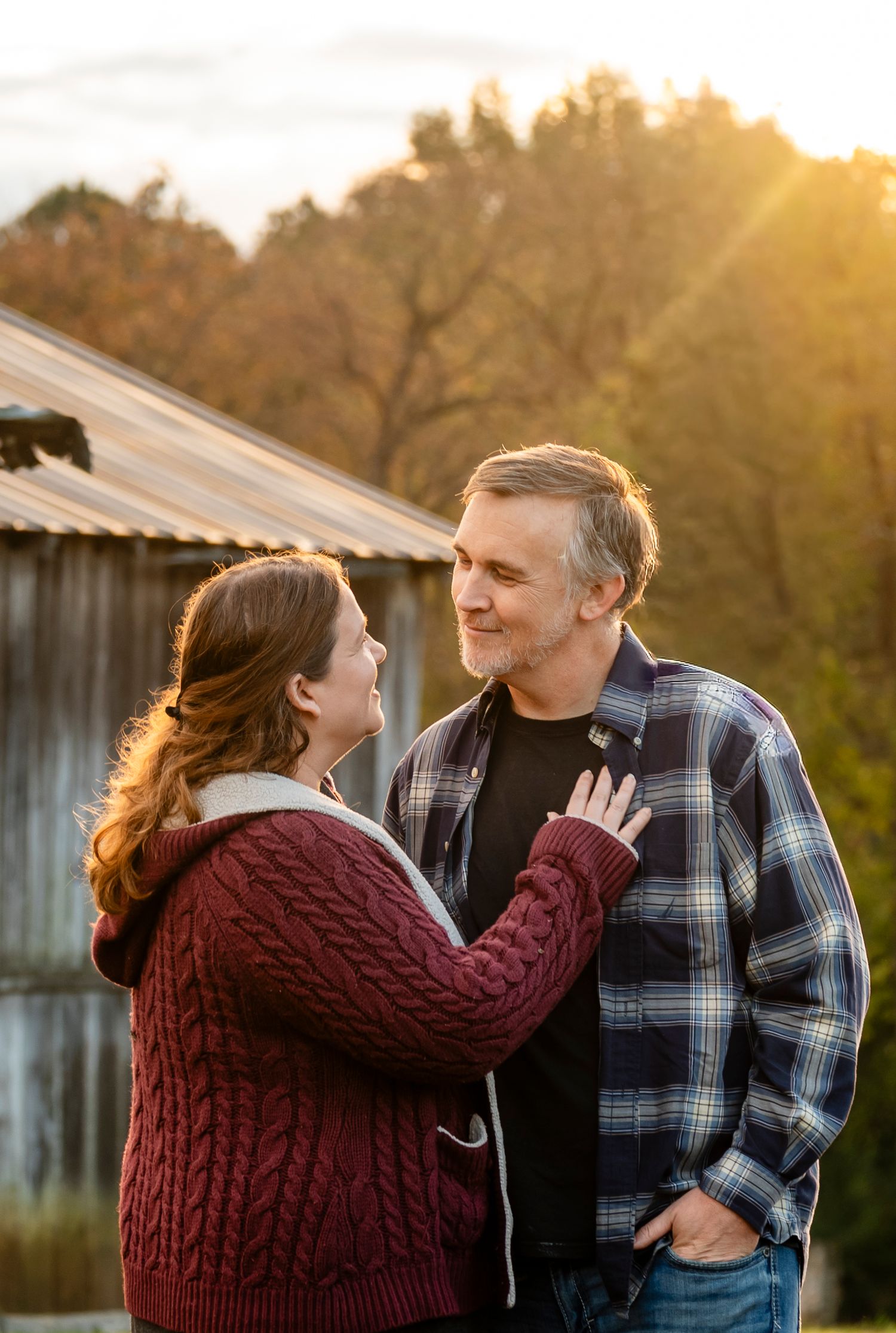 Couple sharing a romantic moment at sunset near a rustic barn, woman in red cable-knit sweater touching mans chest.