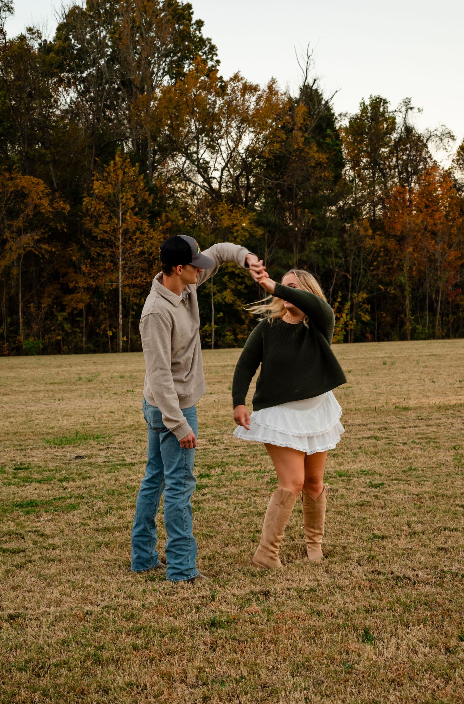 Man spinning woman in white skirt on autumn field at sunset, colorful fall trees in background.