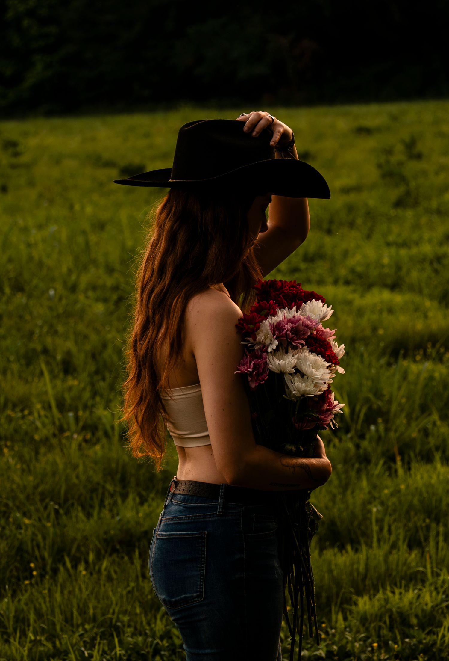 Woman in cowboy hat and jeans holding a bouquet of red, pink, and white flowers in a sunlit field.