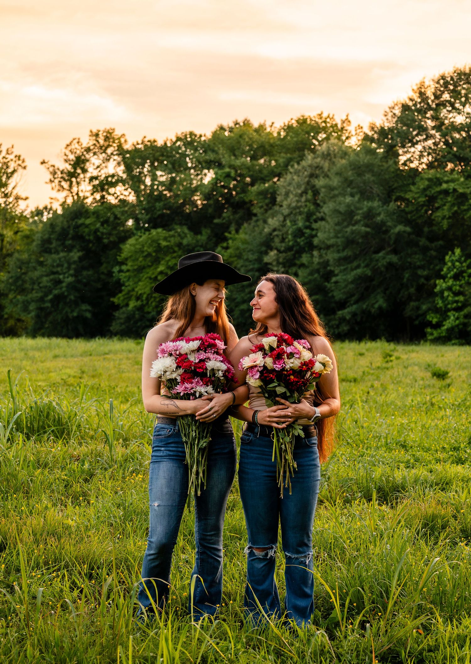 Two smiling women stand in a green field at sunset holding colorful flower bouquets, one wearing a black cowboy hat.