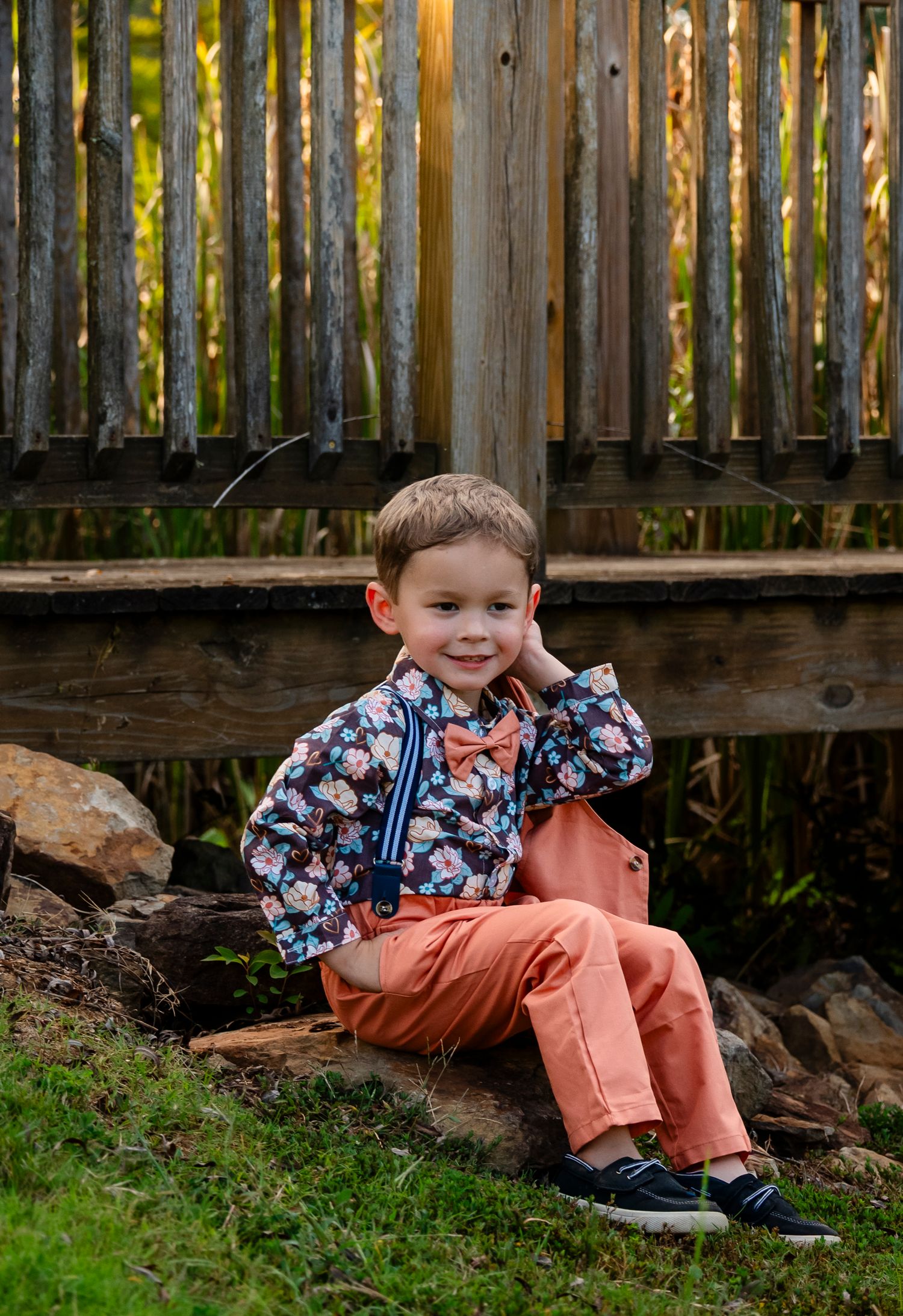 Young boy in floral shirt, orange pants, and bow tie sitting on rocks near a wooden fence outdoors.