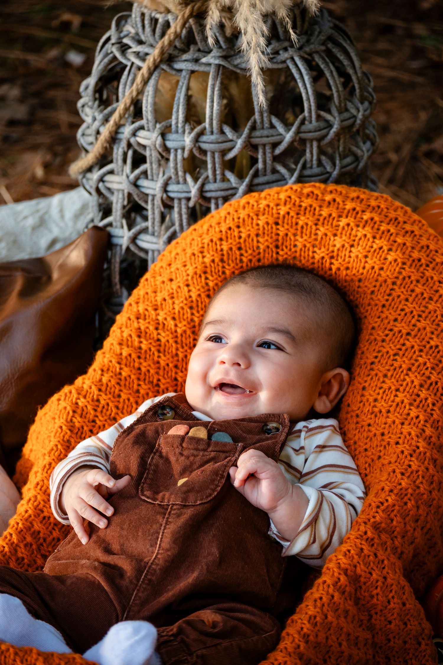 Smiling baby in brown overalls on orange knit blanket with wicker basket in fall setting.