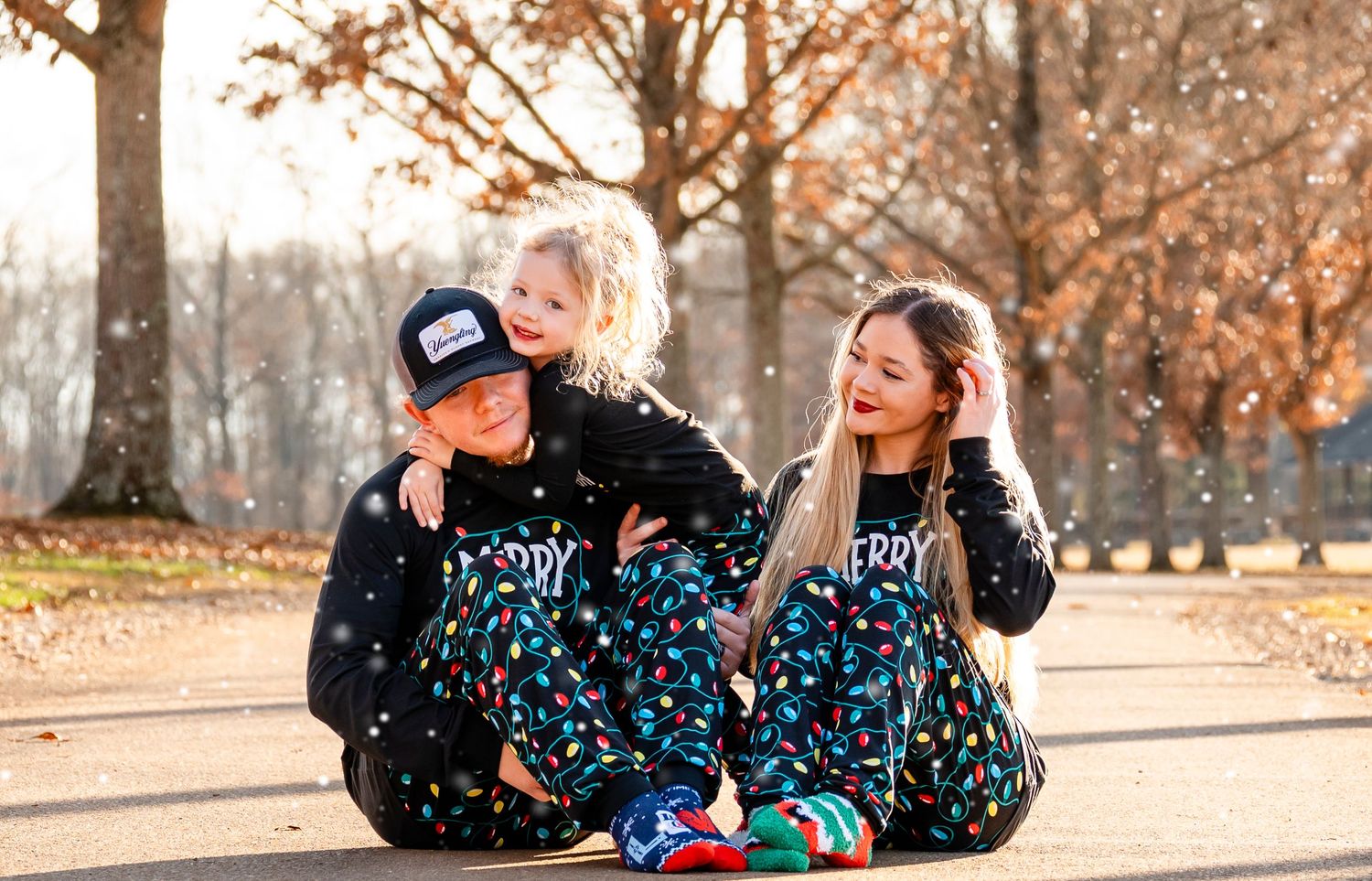 Family of three in matching Merry Christmas pajamas sitting on path with toddler on dad's shoulders.