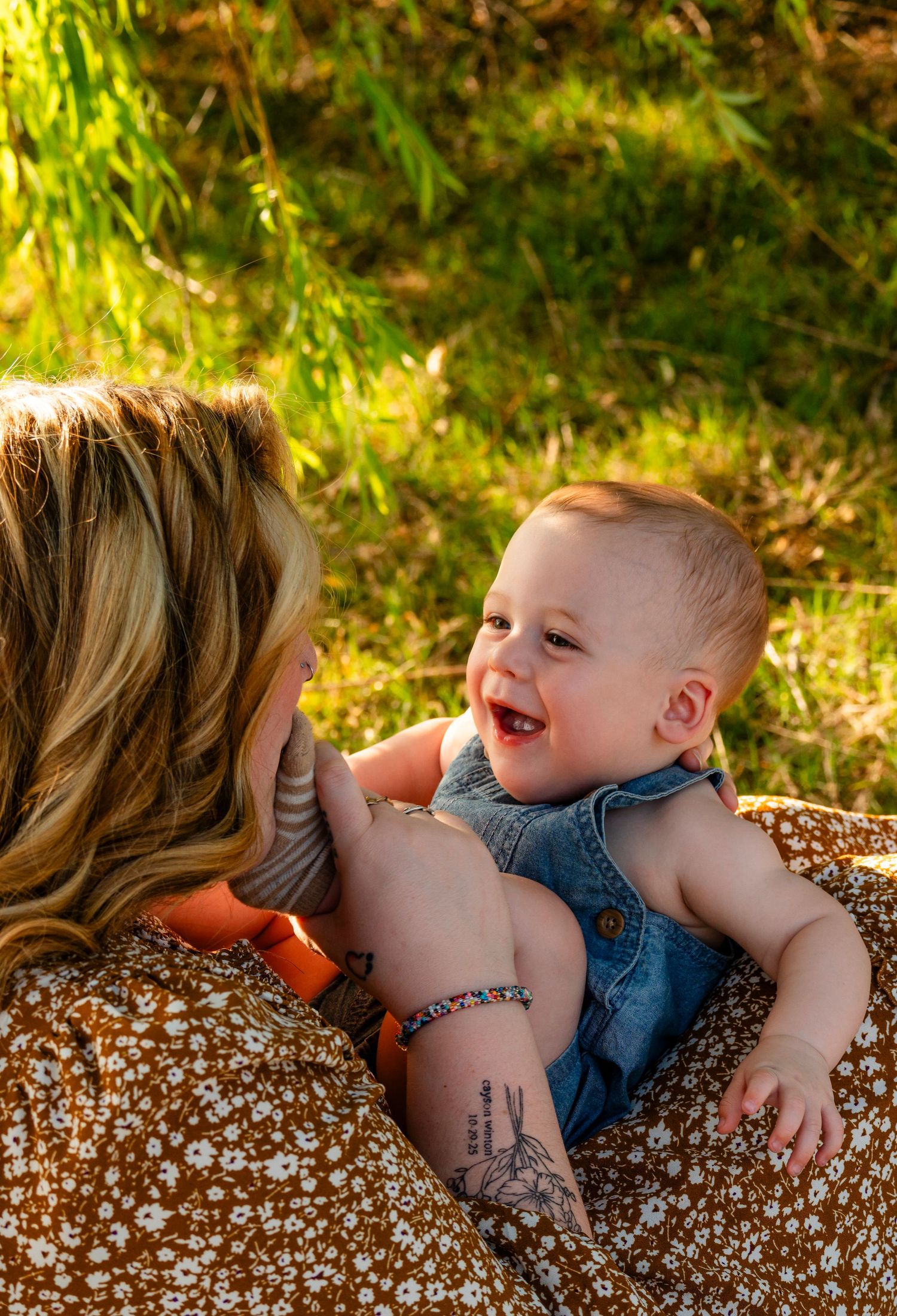 A smiling baby in denim overalls being held by a tattooed mother in a floral dress outdoors.