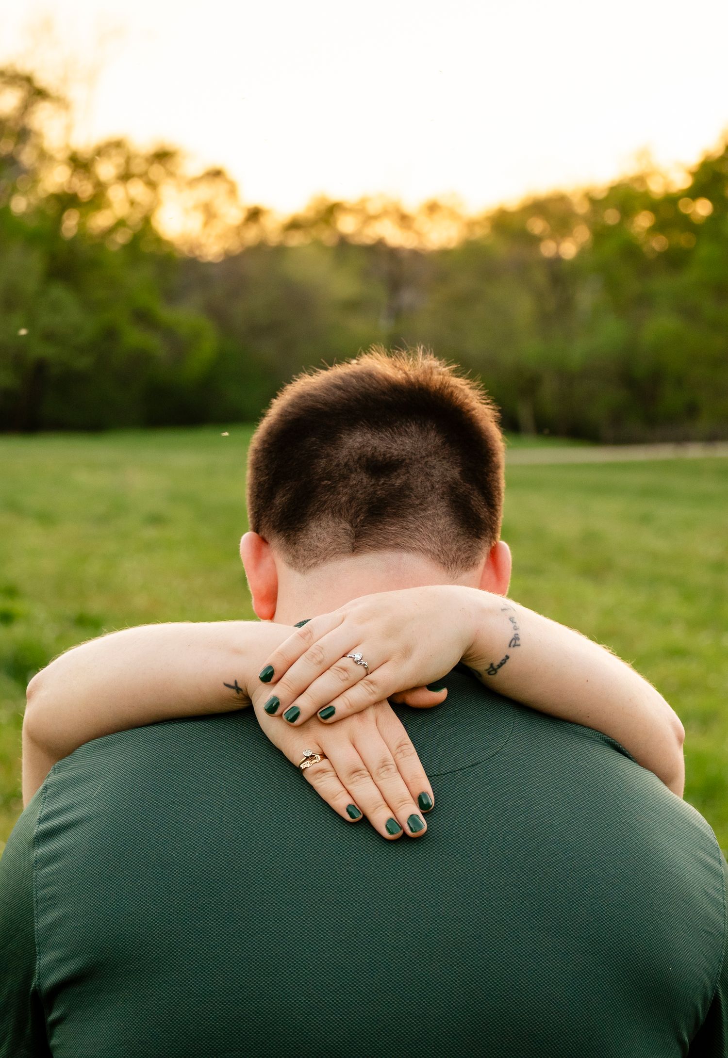 Back view of woman's hands with engagement ring embracing man's neck in a sunny outdoor setting.