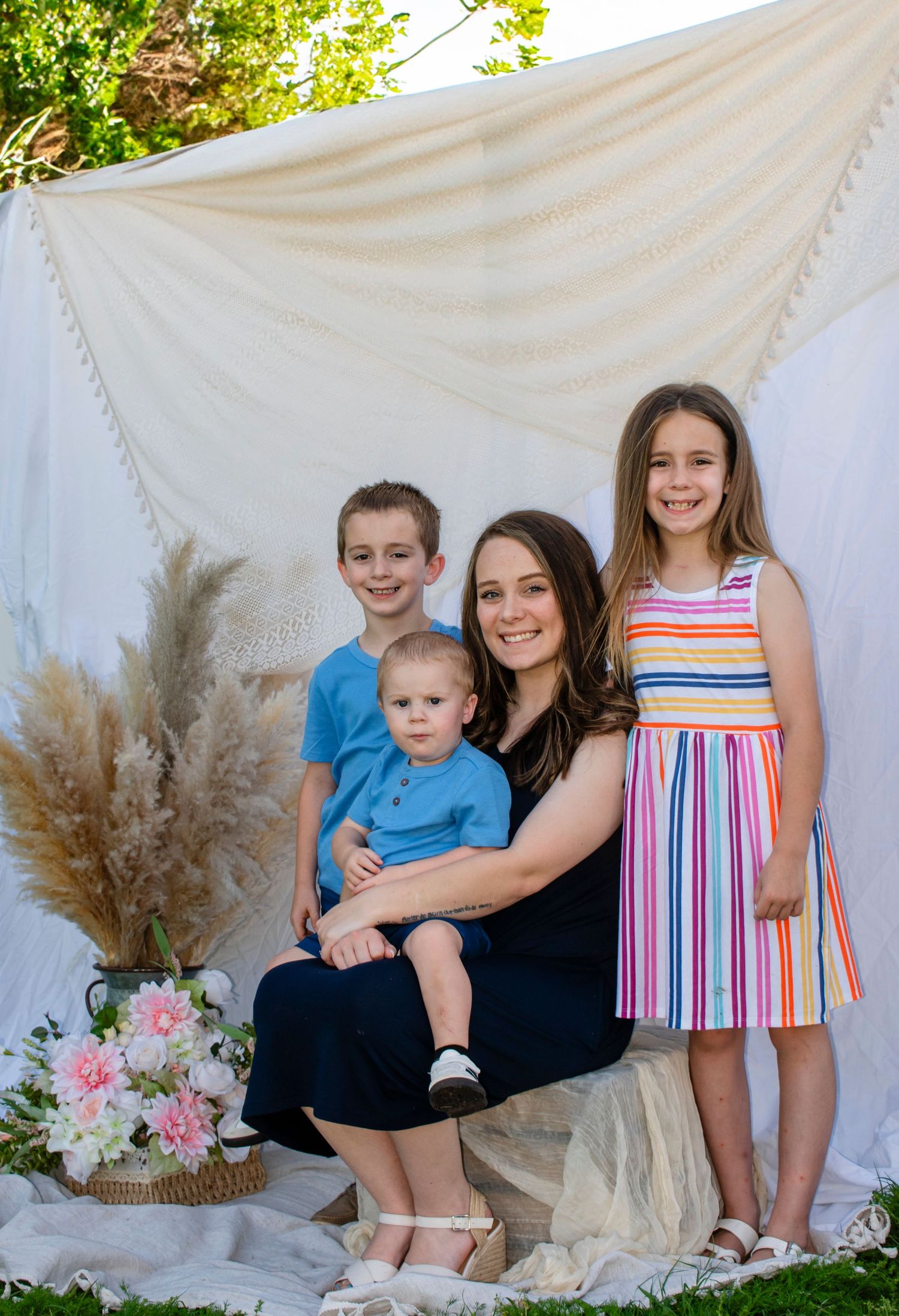 A mother in navy sits with her three children, two boys in blue and a girl in a rainbow striped dress outdoors.