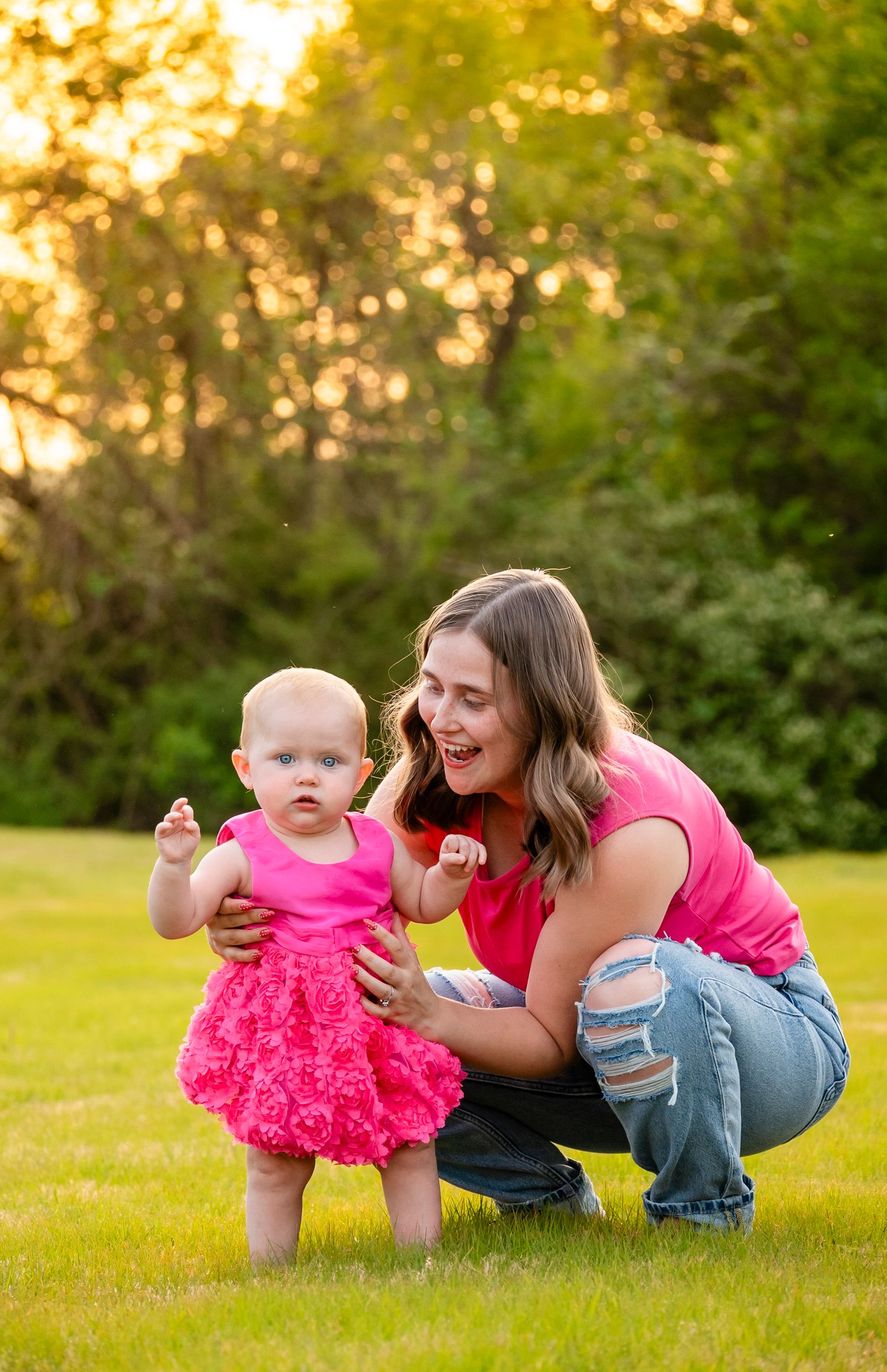 Young mother in pink top helps baby girl in pink dress take first steps on green lawn.