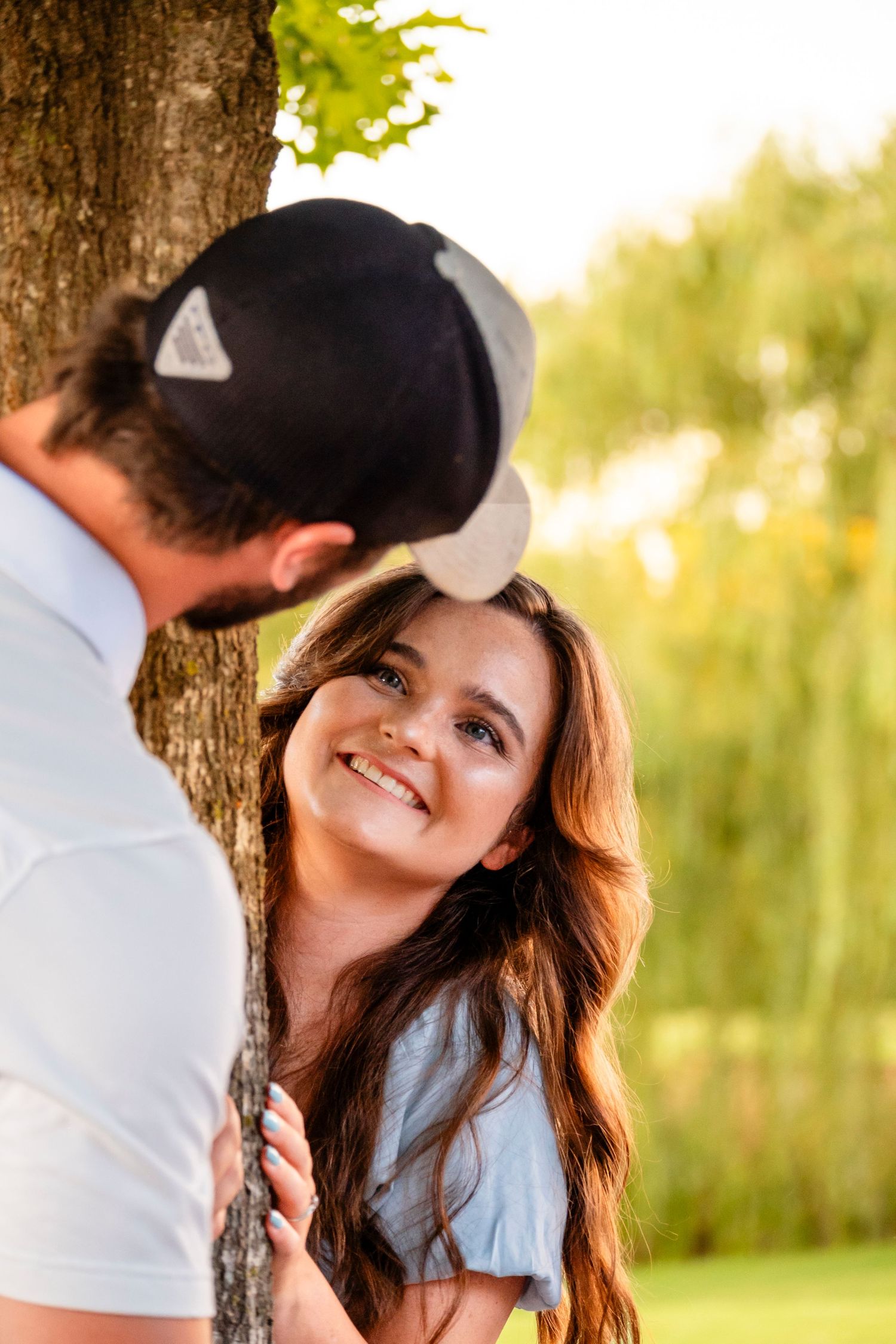 A smiling young woman leans against a tree looking up at her partner during an outdoor couples session.
